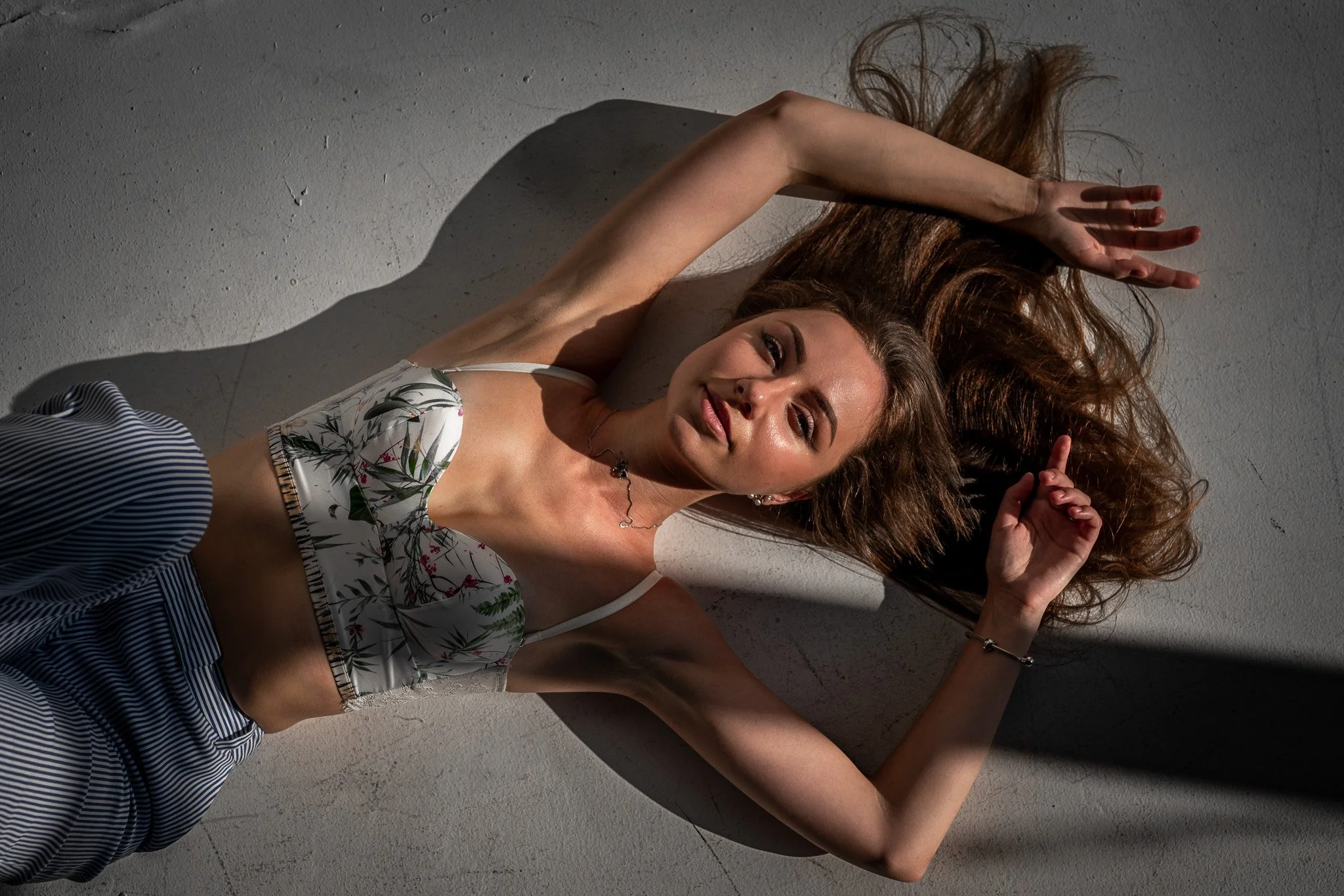 A young woman with long brown hair lying on a gray floor, casting a shadow from a strong overhead light, wearing a floral crop top and striped pants, looking at the camera.