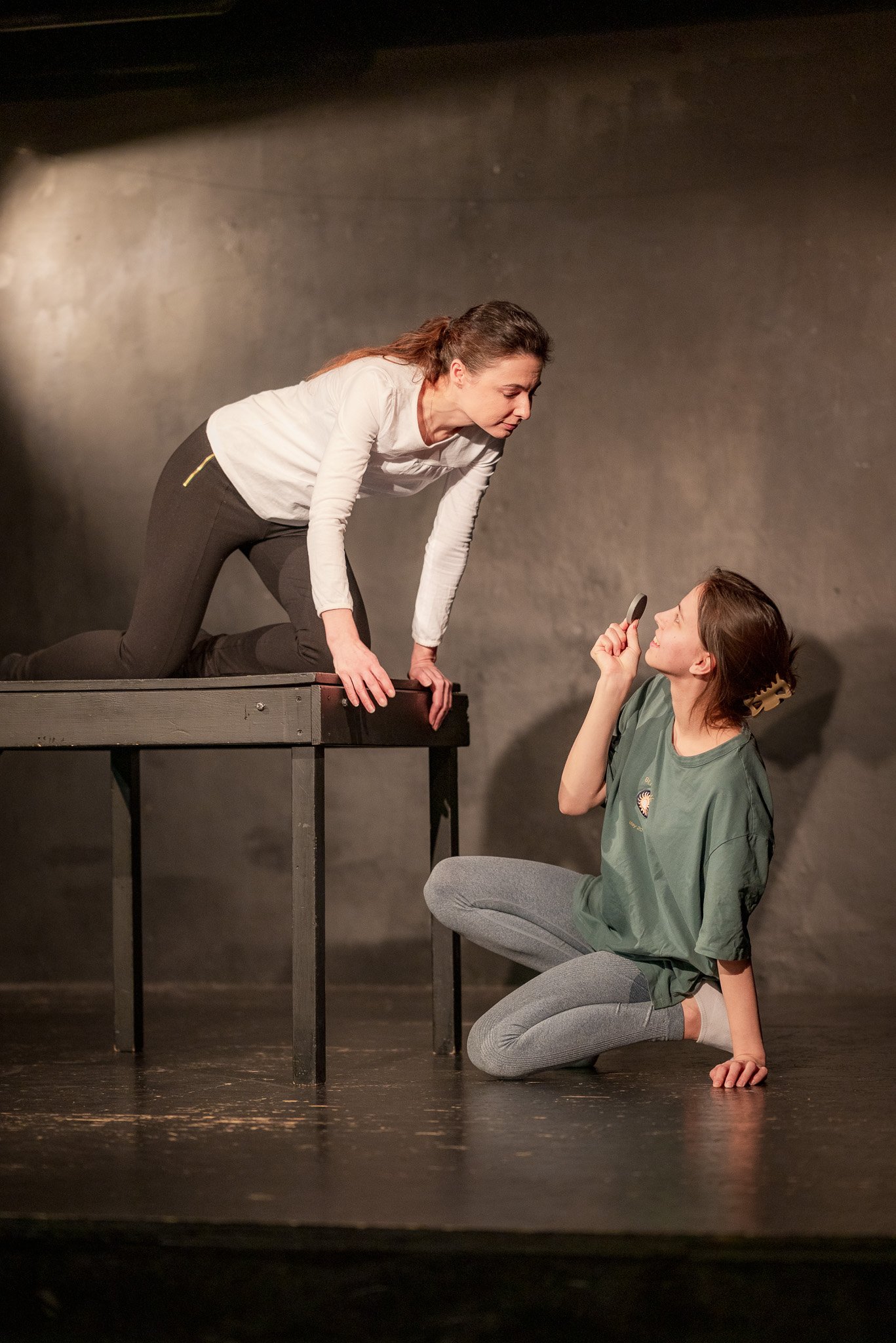 Two women performing a theatrical scene, one kneeling with a prop resembling a magnifying glass, the other leaning forward on a wooden platform, in a dimly lit room.