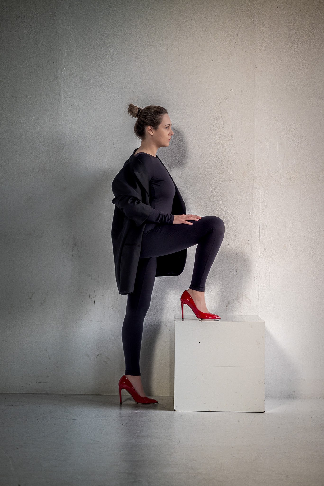 Woman in black outfit with red high heels, one foot on a white block, side profile view.