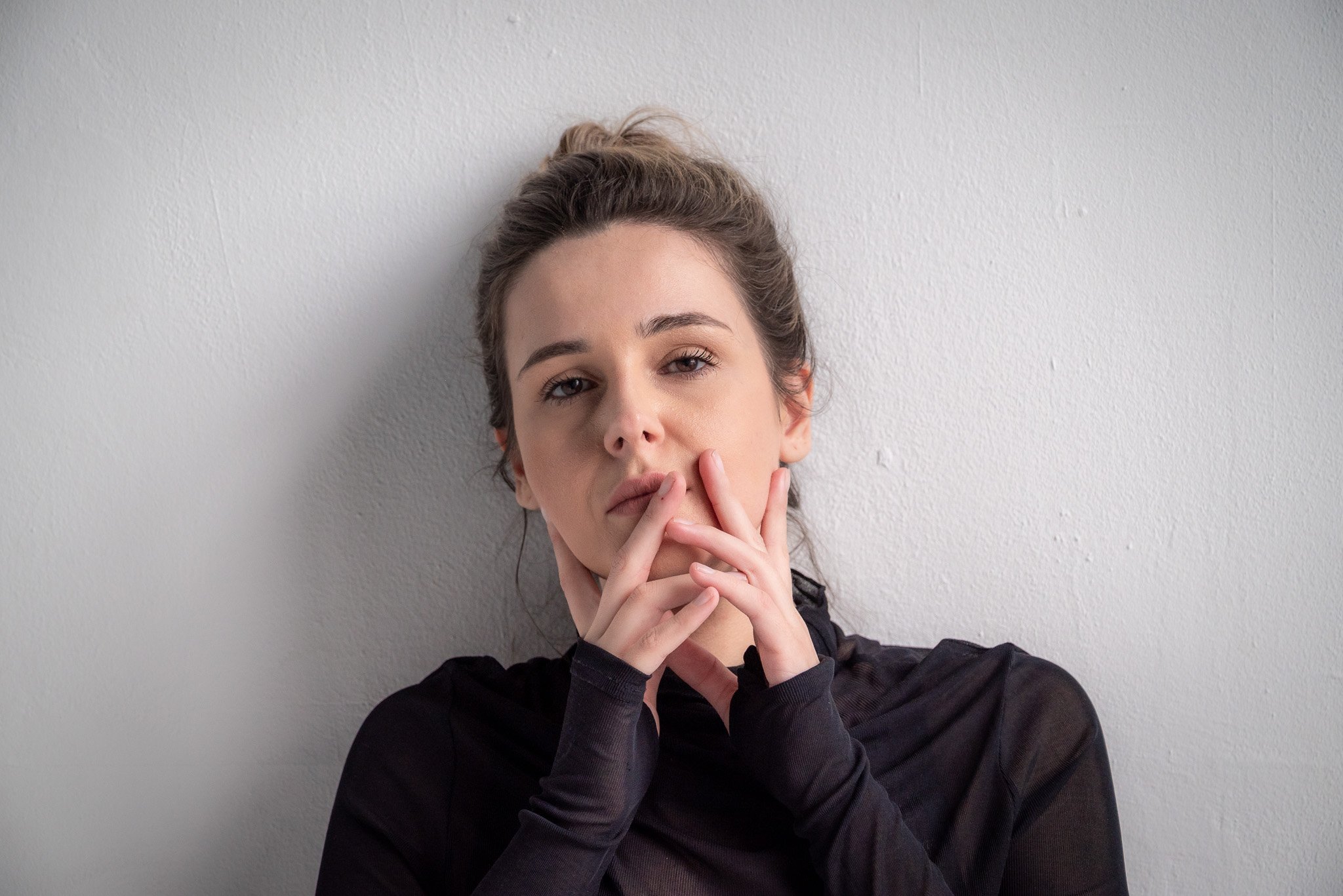 Woman with hands near face, wearing a black shirt, posing against a white wall.