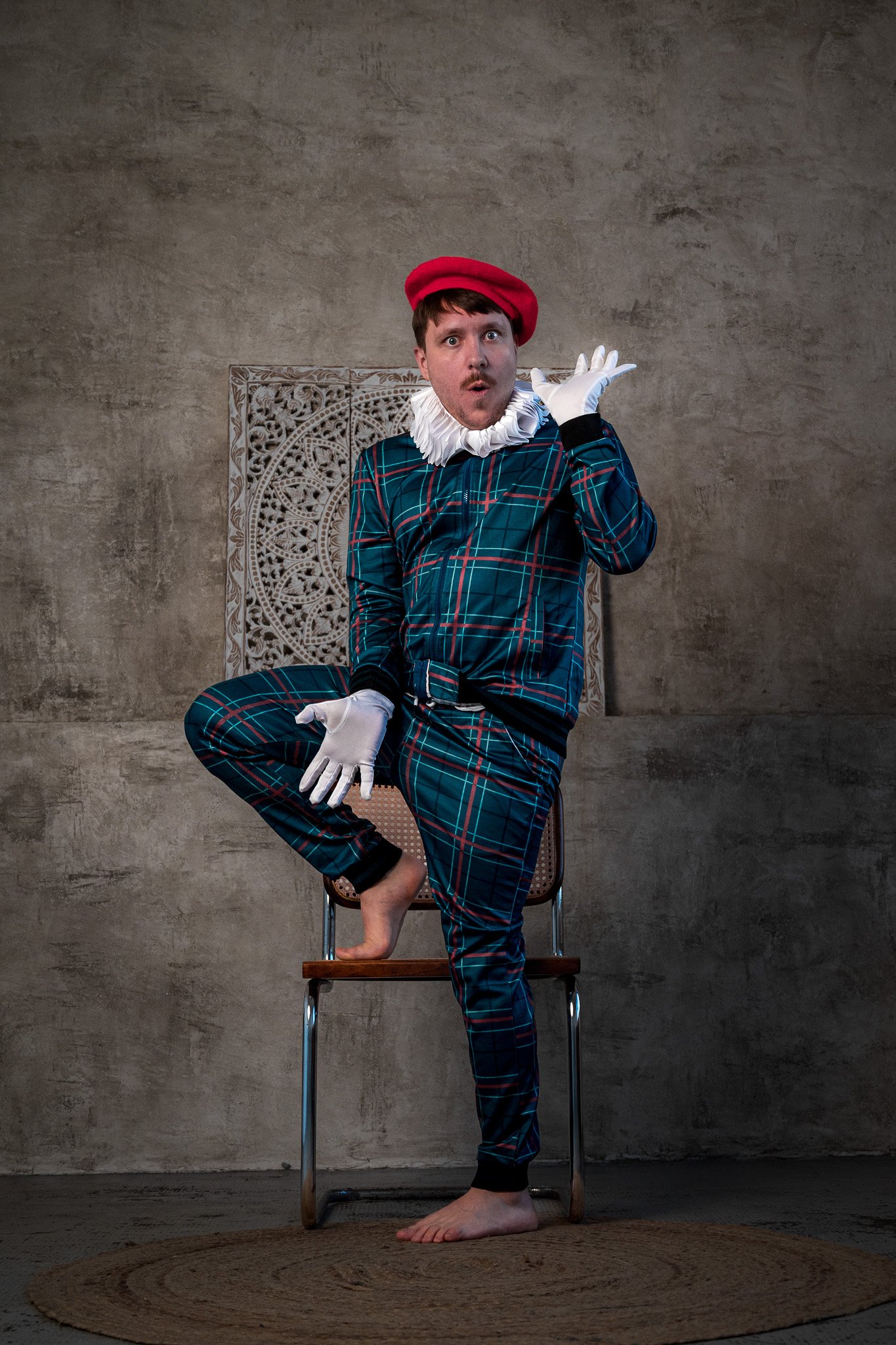 Person in plaid jumpsuit, red beret, white gloves, and ruffled collar posing on a chair.