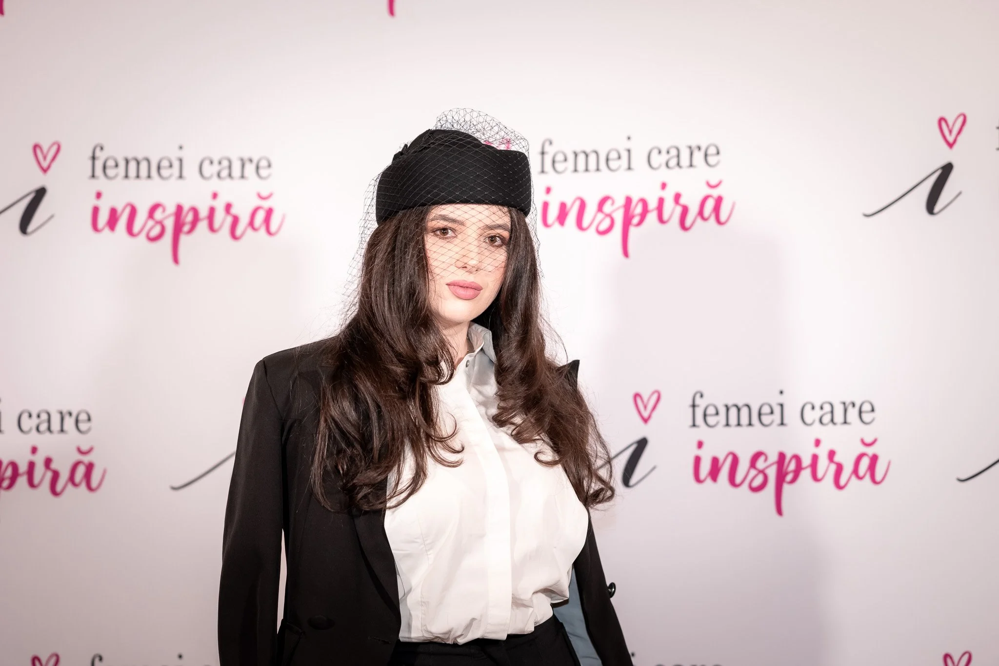 A woman with long, wavy brown hair, wearing a white blouse, a black blazer, and a black hat with a mesh veil, standing in front of a white background with pink and black text that reads "femei care inspiră."