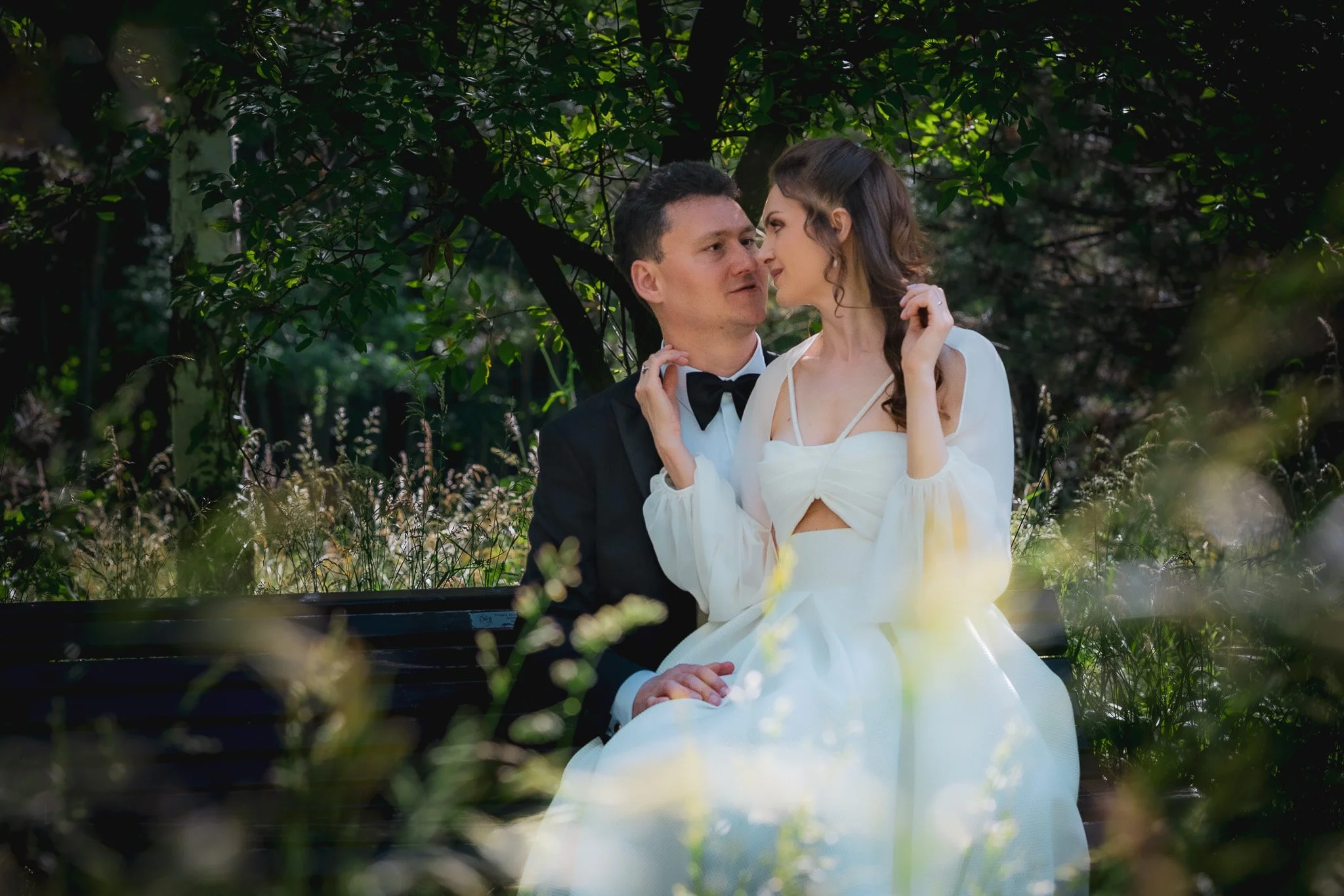 A couple in formal attire sitting on a bench in a lush green park, gazing into each other's eyes.