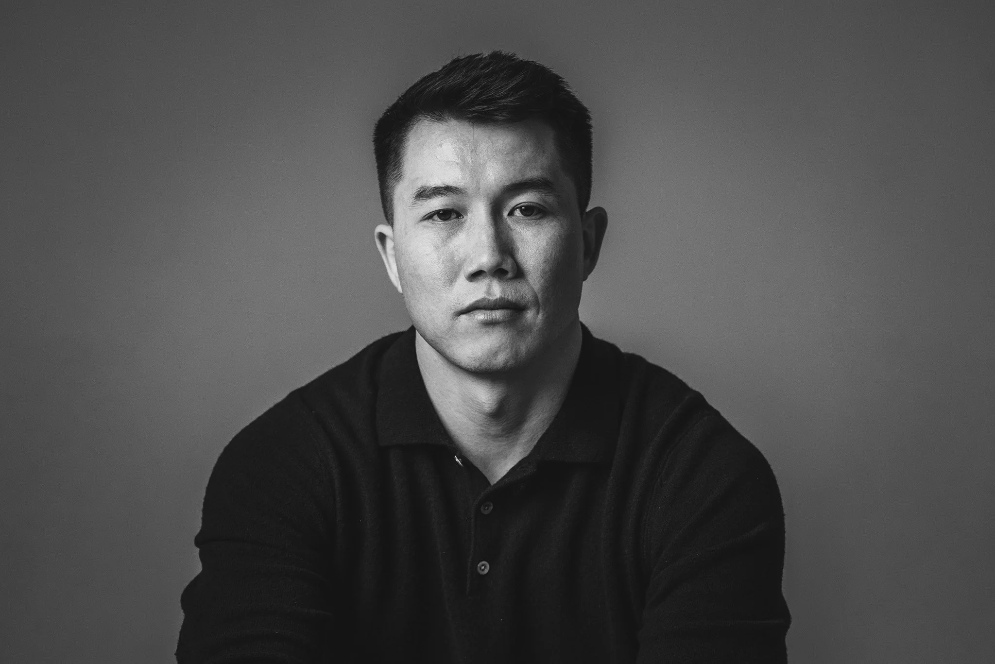 Black and white portrait of a young man with short dark hair, wearing a black collared shirt, looking directly at the camera with a serious expression.