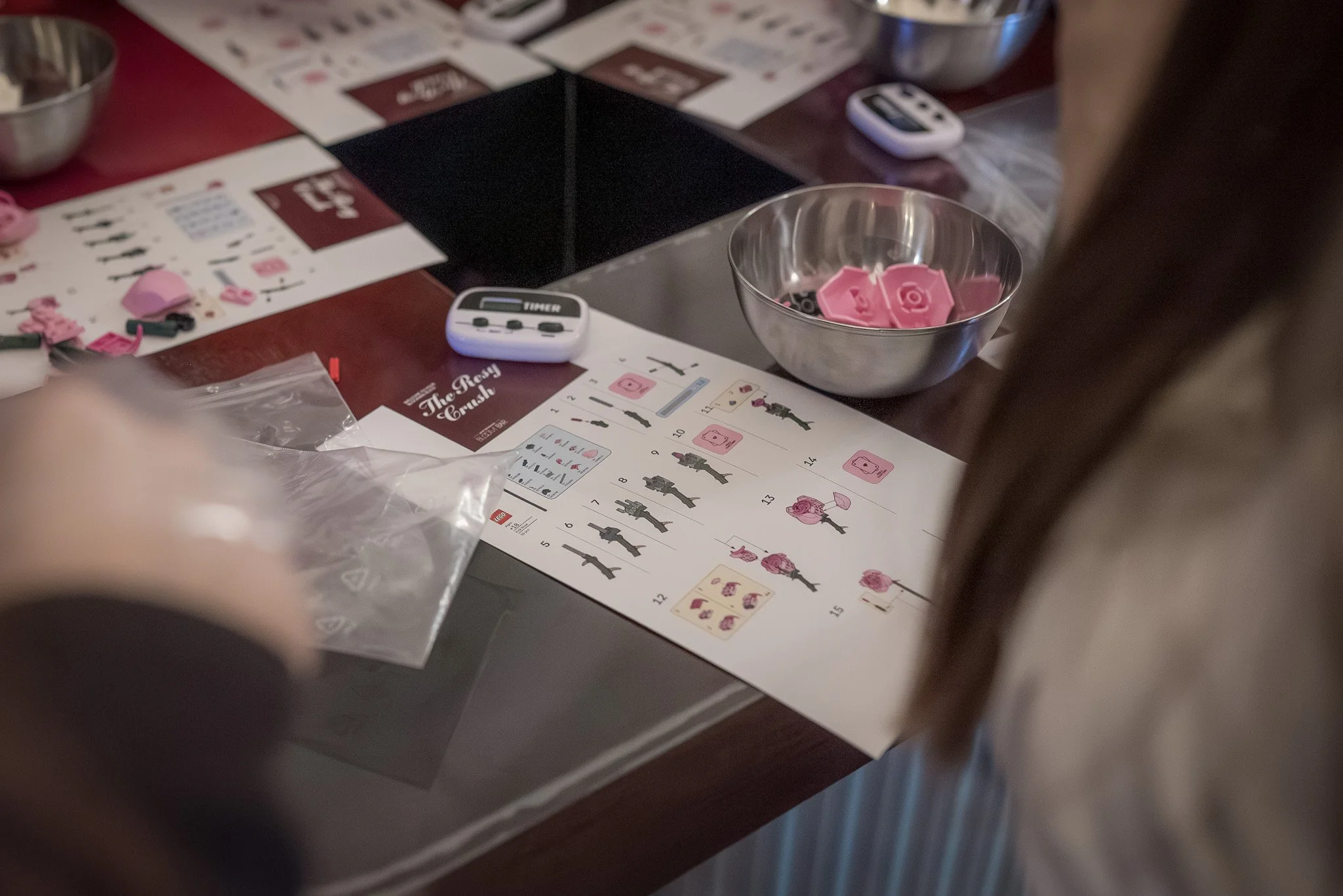 A close-up of a table with plastic flower-making kits, pink flower parts, instruction sheets, a remote control, and bowls, with a person partially visible.