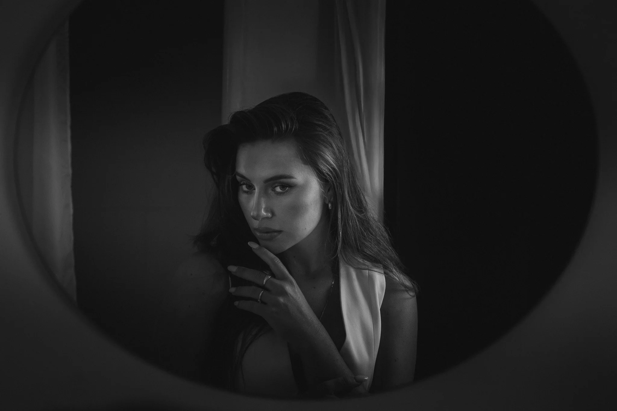 Černobílá fotografie ženy s dlouhými vlasy, která se dívá do zrcadla s prstem dotýkajícím se rtů, odráží se v kruhovém rámu zrcadla.