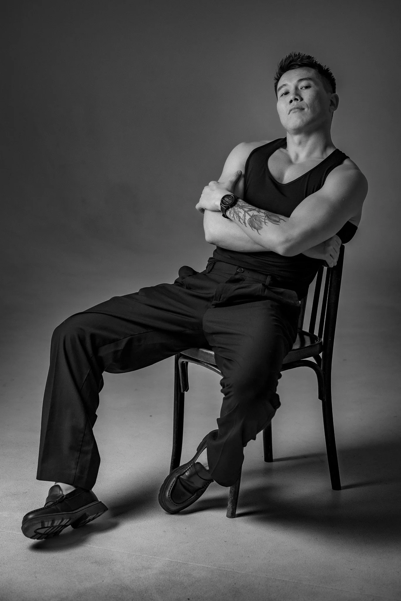Black and white photo of a young man sitting on a chair with crossed arms, wearing a sleeveless top, pants, and shoes, with a tattoo on his left arm, against a plain background.