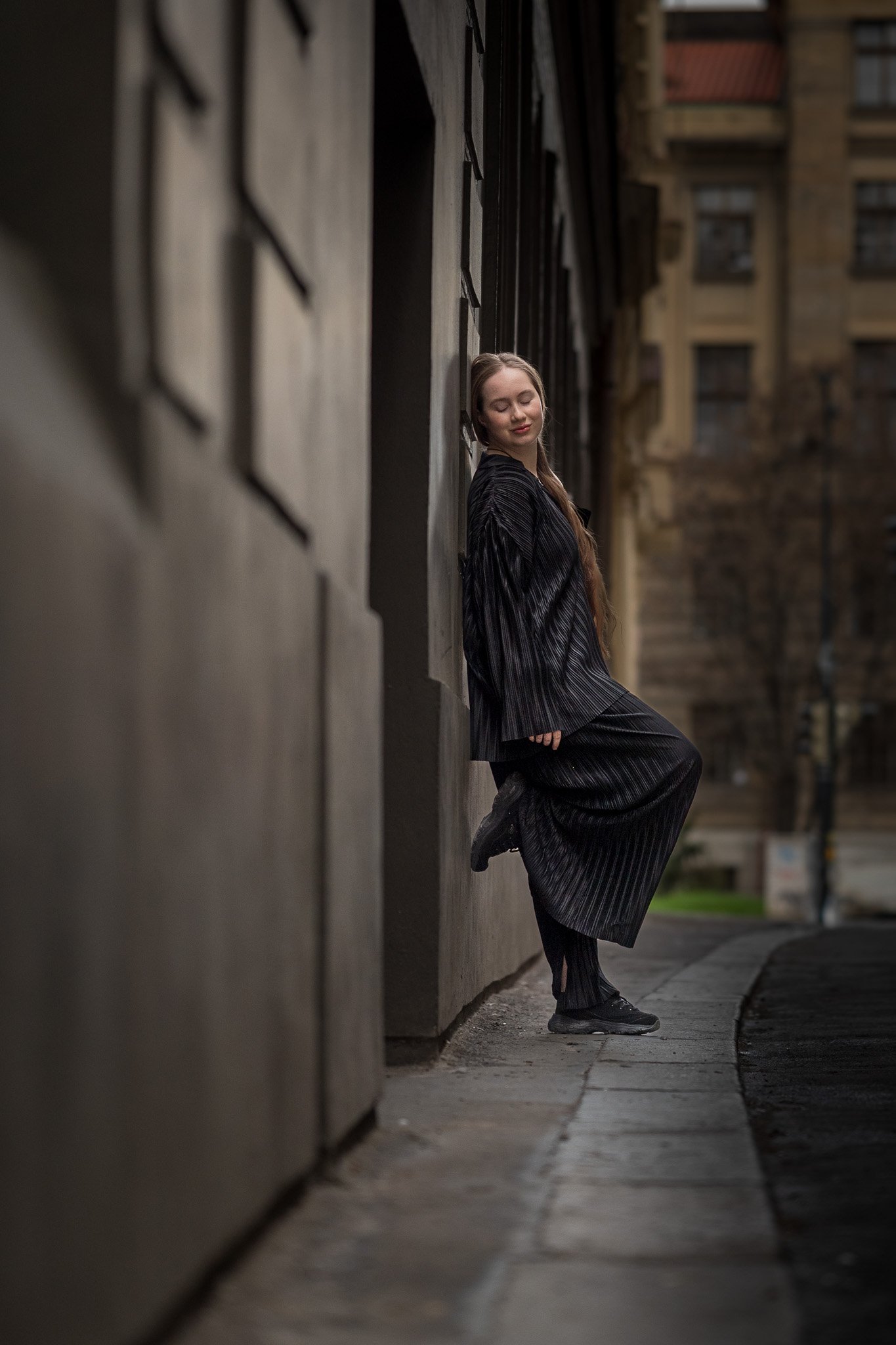 Woman in black outfit leaning against a building on a city sidewalk.