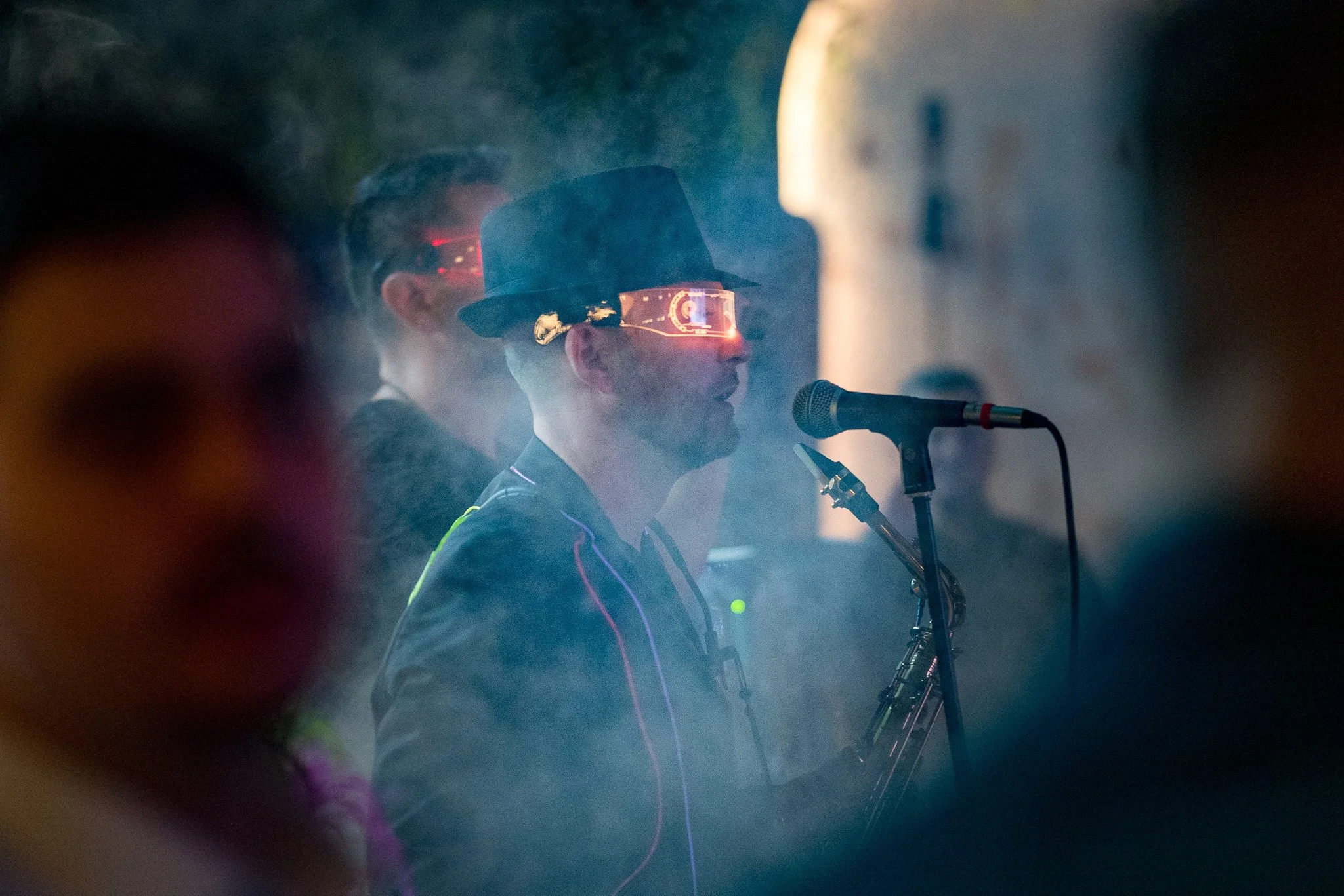 A person wearing futuristic glasses and a hat speaking into a microphone, surrounded by smoke with people in the background.