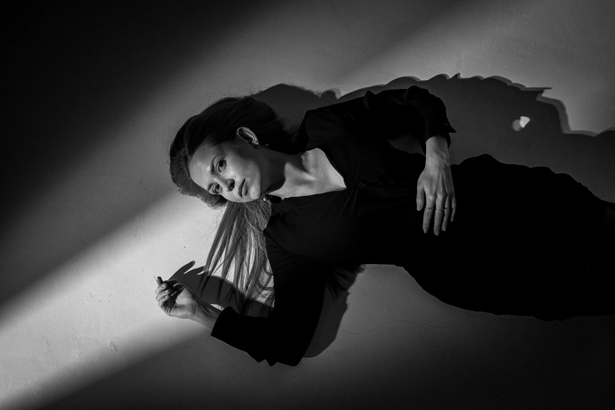Black and white photo of a woman lying on the floor, casting a long shadow on the wall, holding a cigarette, with her head turned slightly to the side and her hair spread out.
