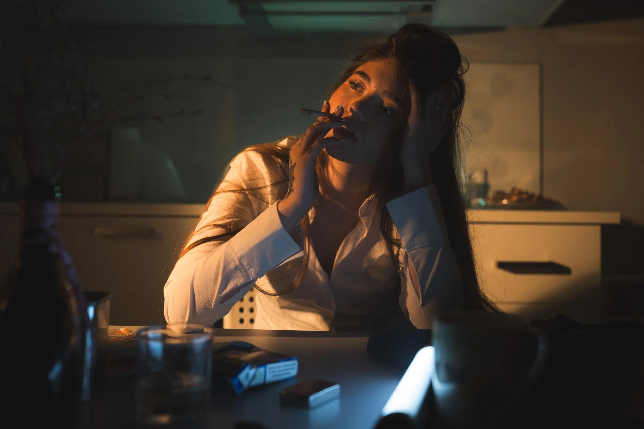 A photo of a woman in Prague with long hair, wearing a white shirt, is sitting at a table smoking a cigarette in a dimly lit room.