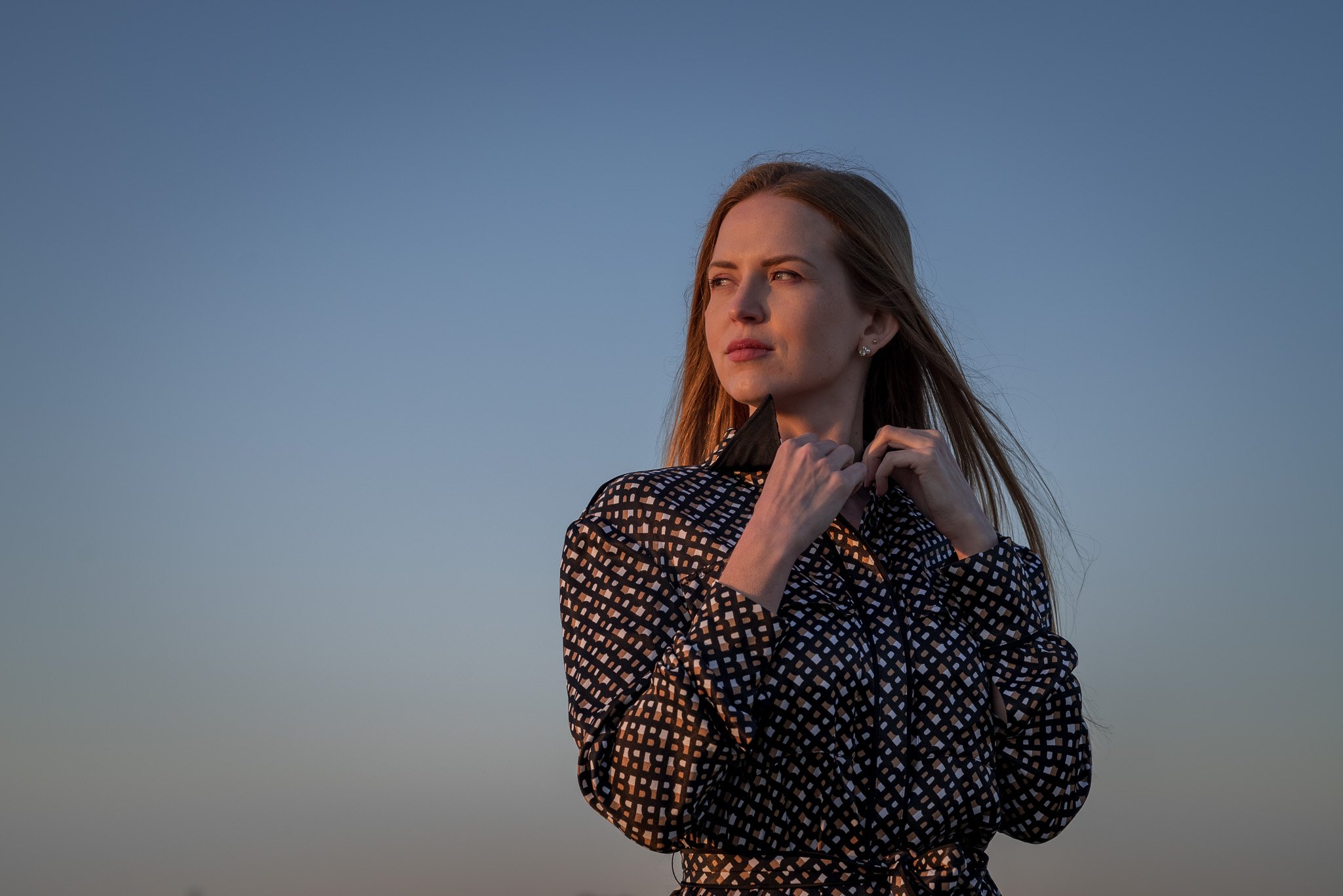 Woman in patterned coat standing outdoors during golden hour with a clear sky background.