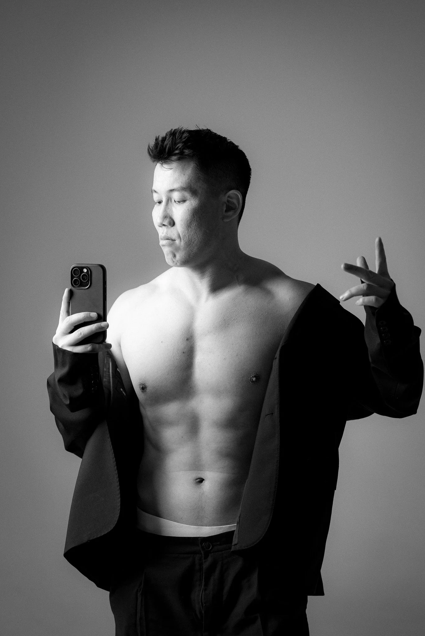 Black and white photo of a shirtless man taking a mirror selfie with a smartphone, partially wearing an unbuttoned black jacket.