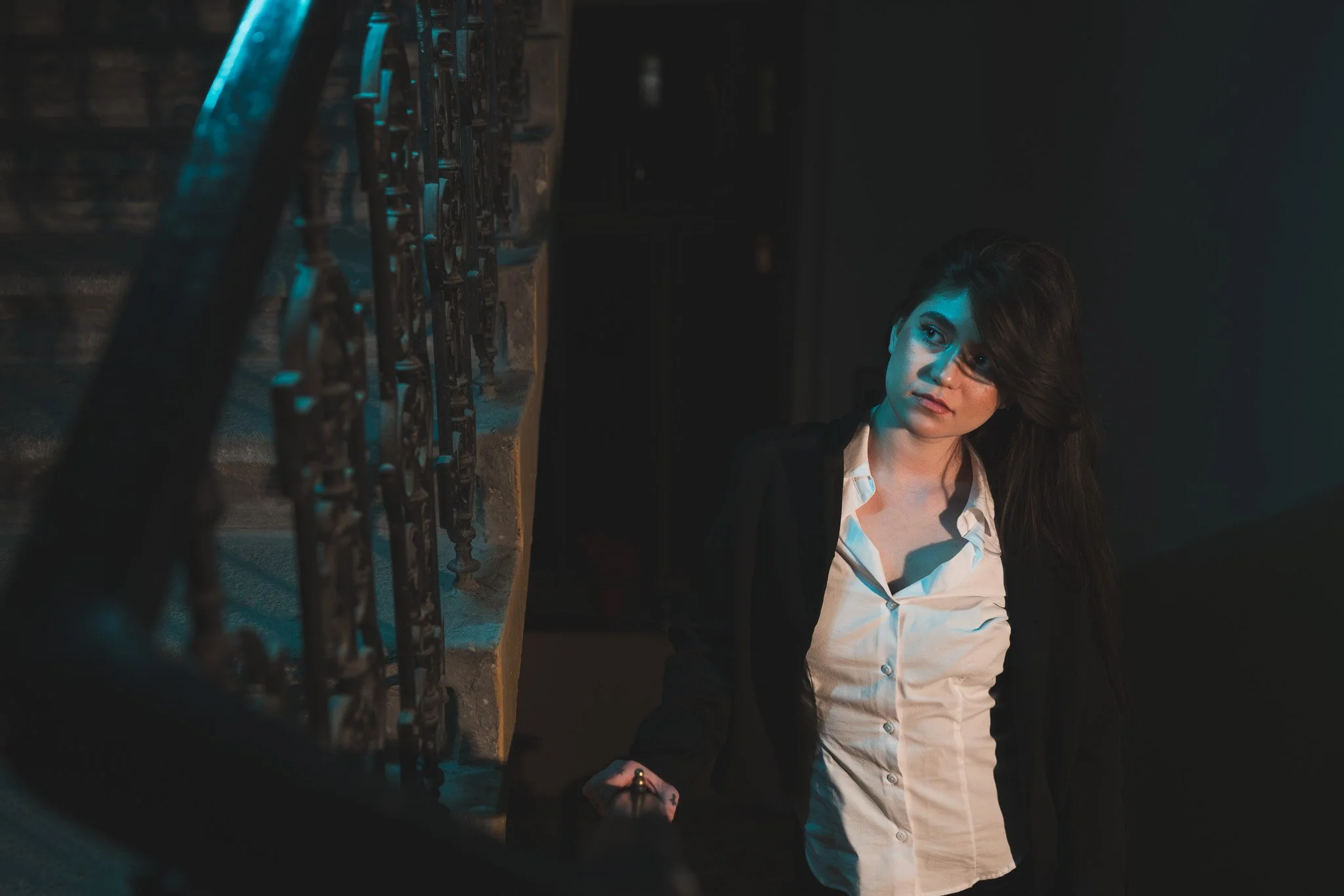 A woman with dark hair, wearing a white shirt and black blazer, standing near a staircase with ornate iron railings, illuminated by moody blue lighting.