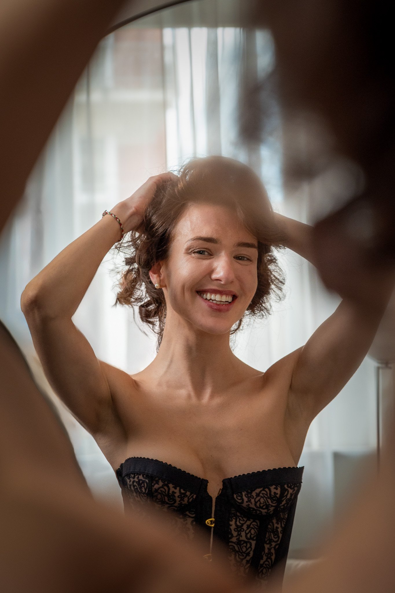 Smiling woman touching her hair, wearing a black lace bustier, looks in mirror.