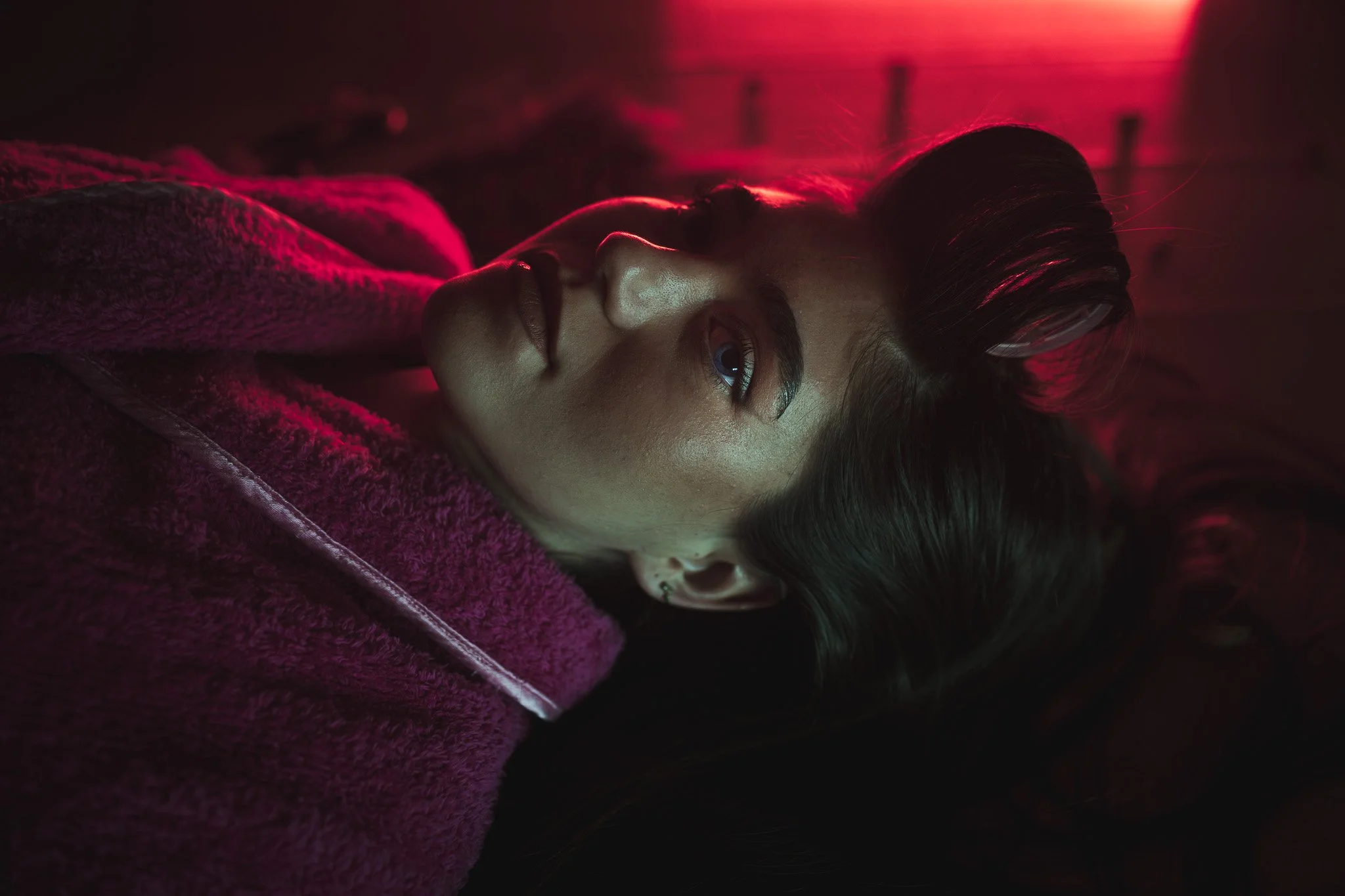 A portrait photograph of a woman with dark hair and blue eyes lying down, wearing a dark pink and purple fuzzy coat, with red lighting illuminating the background. Taken in Prague.