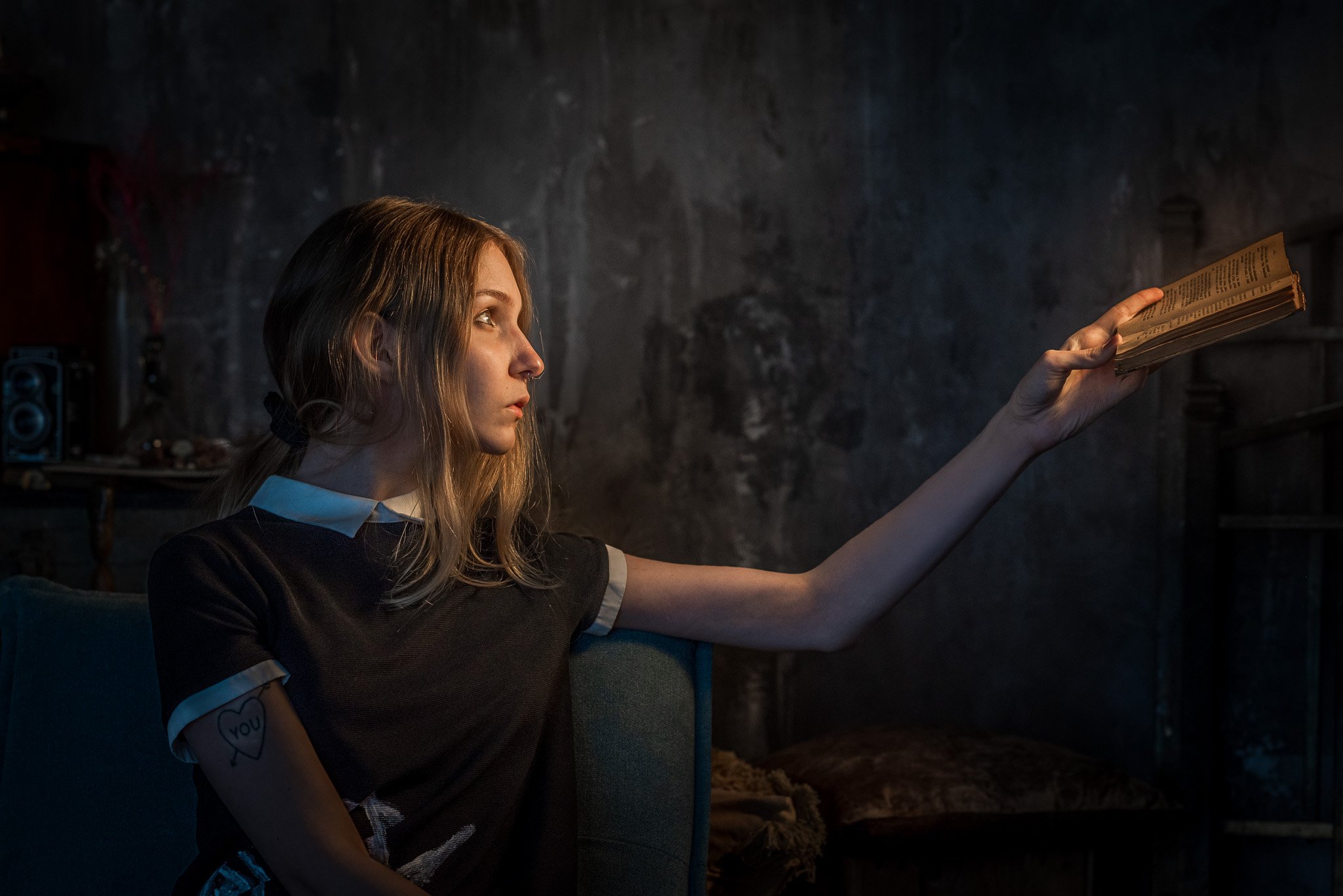 A young woman with blonde hair, dressed in a black shirt with white trim, sitting in a dimly lit room, holding a book with her arm extended.
