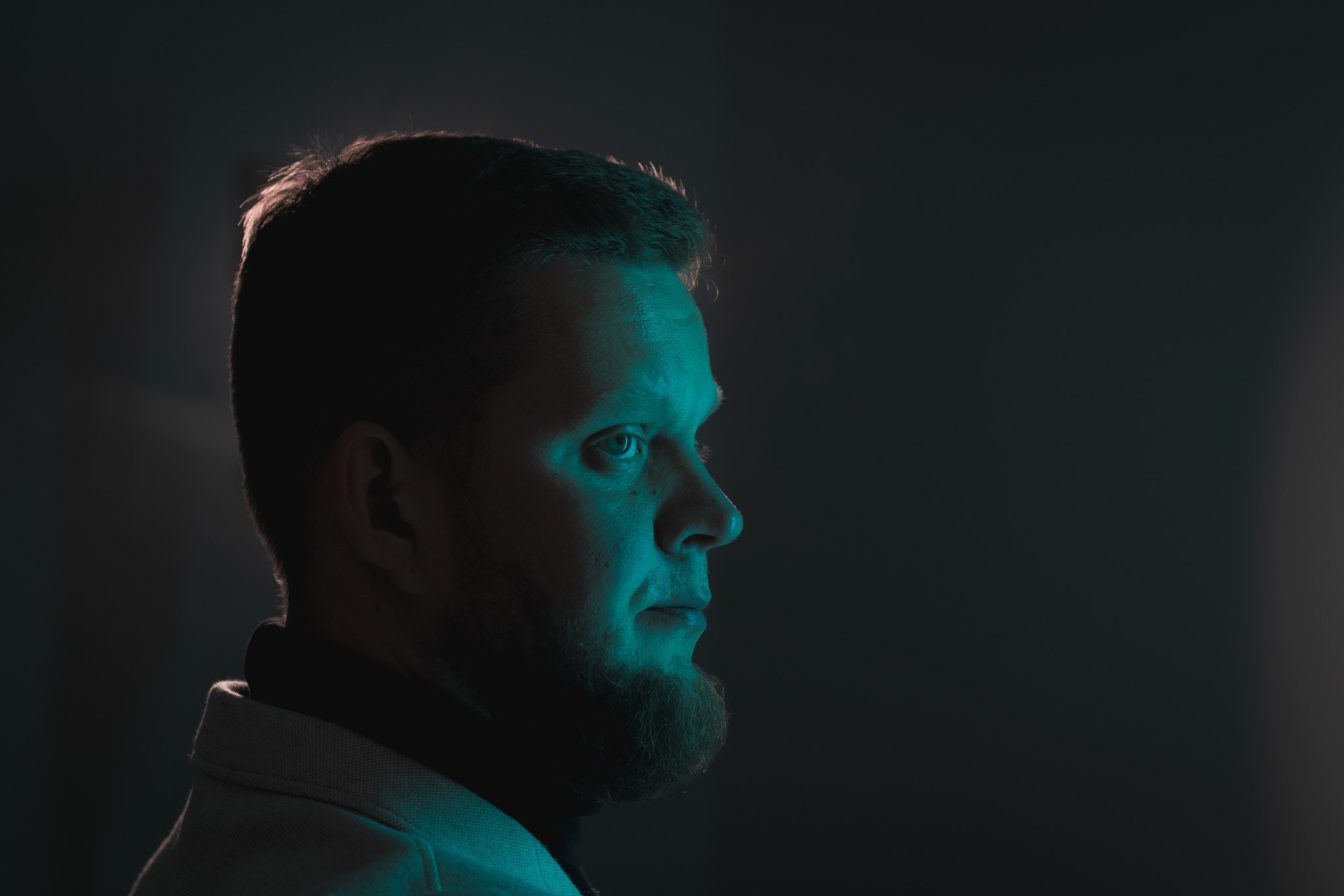 Boční profil muže s krátkými vlasy a plnovousem, osvětlený tyrkysovým a teplým světlem, na tmavém pozadí.