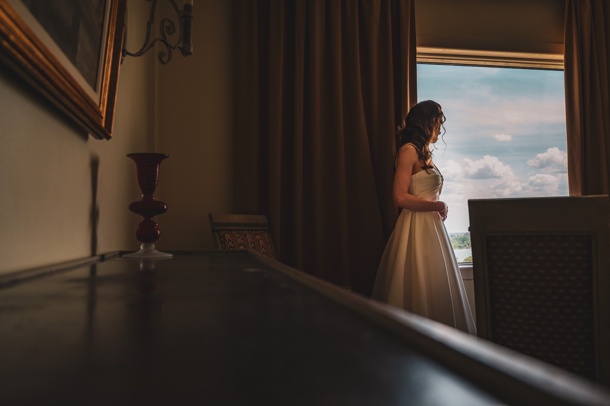 Woman in wedding dress standing by window with brown curtains; room with dark furniture and red vase