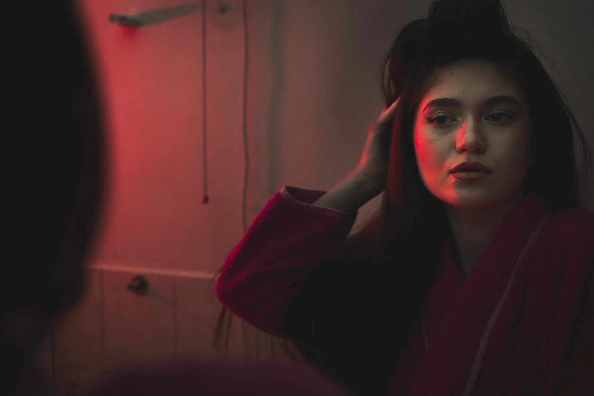 A photo of a young woman with dark hair and makeup looking at her reflection in a mirror, with red and green lighting creating a moody atmosphere.