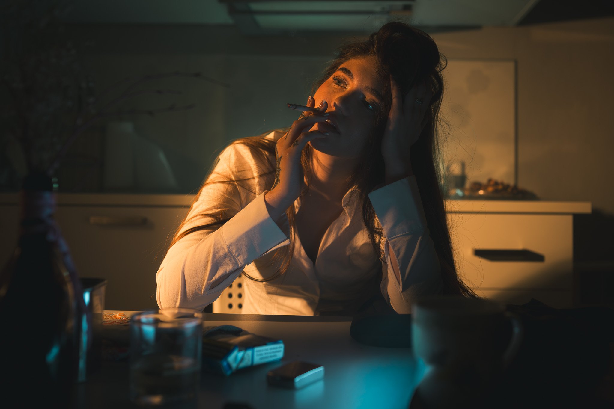 Photo of a woman with long dark hair sitting at a table in a dimly lit room, smoking a cigarette, with her hand on her forehead, wearing a white shirt.