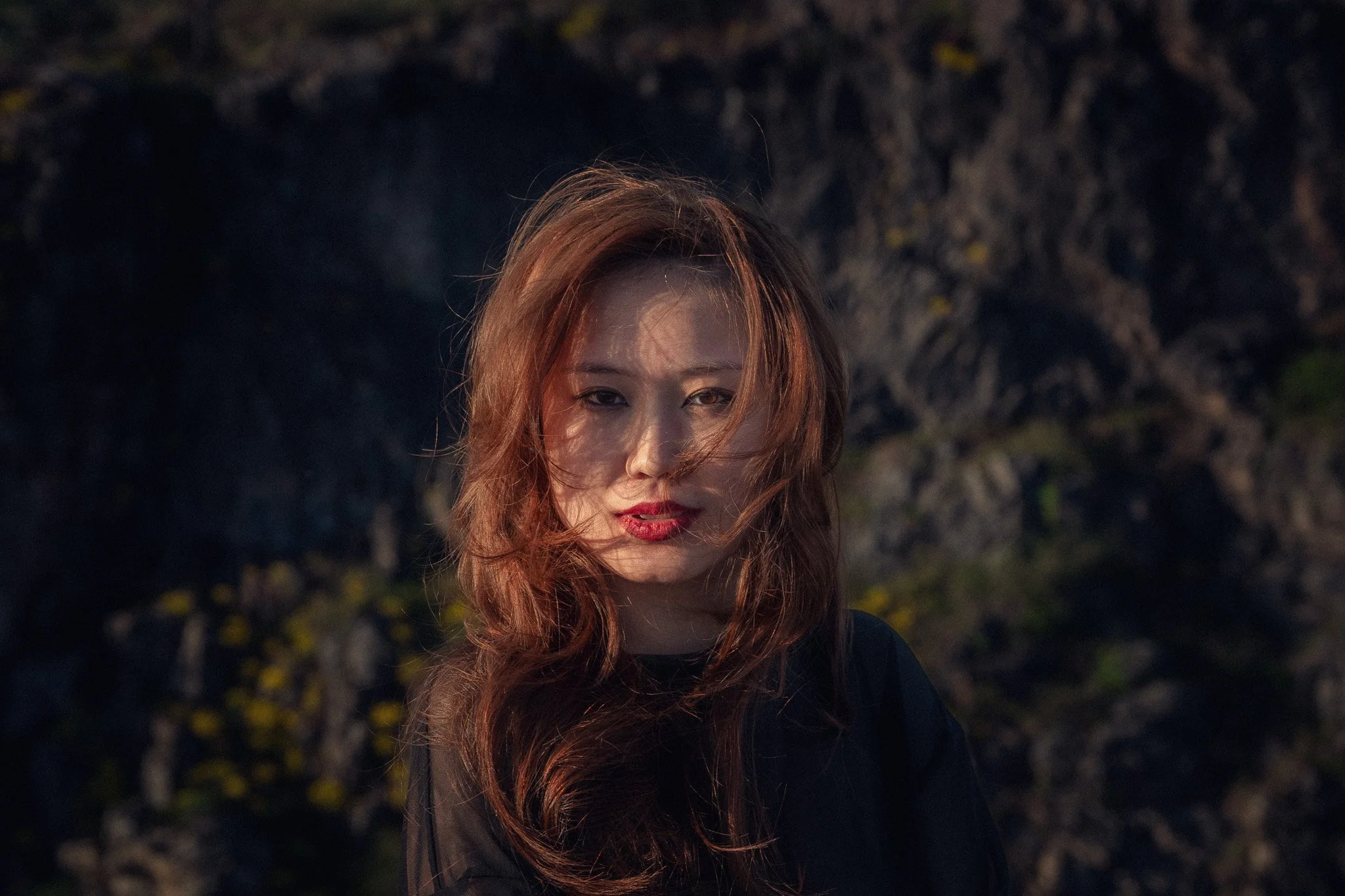 Woman with long, wavy reddish-brown hair, wearing dark clothing, standing outdoors with a rocky background, highlighted by warm sunlight.