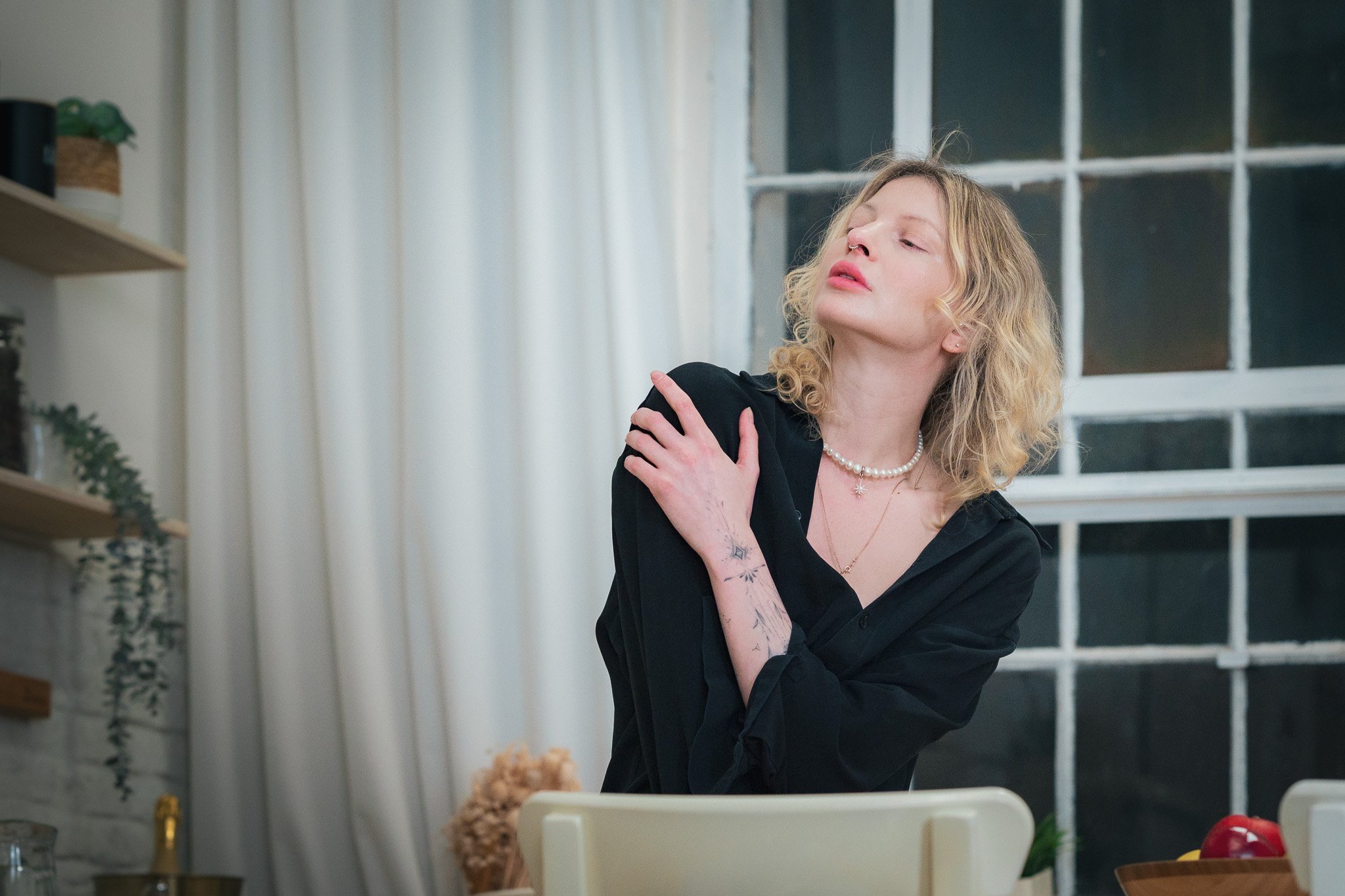 Person with blond curly hair, wearing a black shirt and pearl necklace, posing indoors near a window.