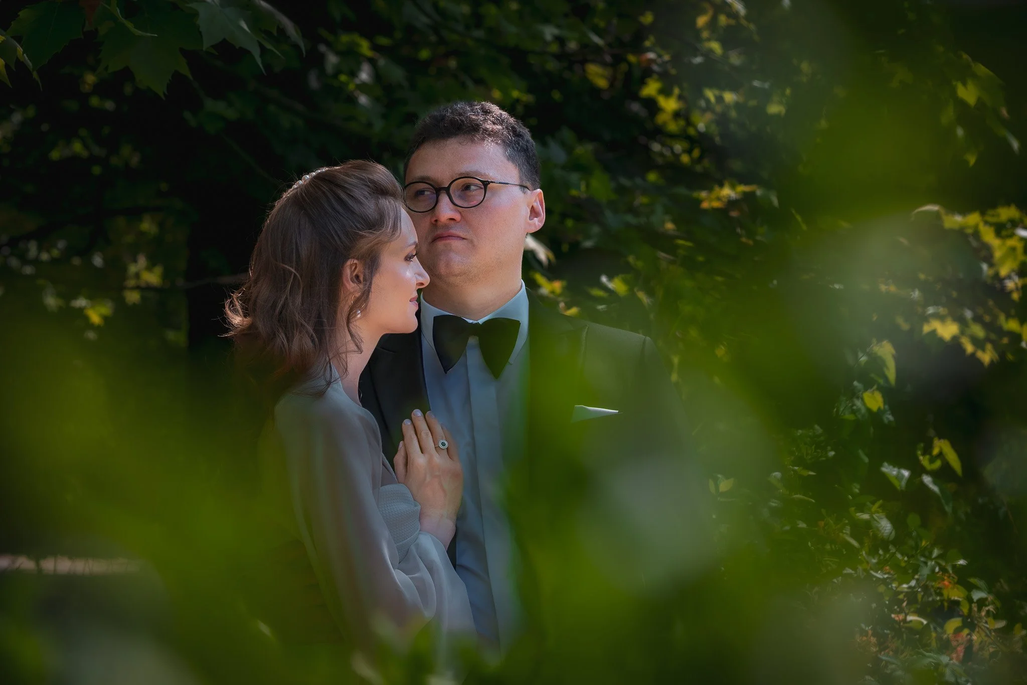 A married couple in formal attire sharing an intimate moment outdoors with green foliage surrounding them.