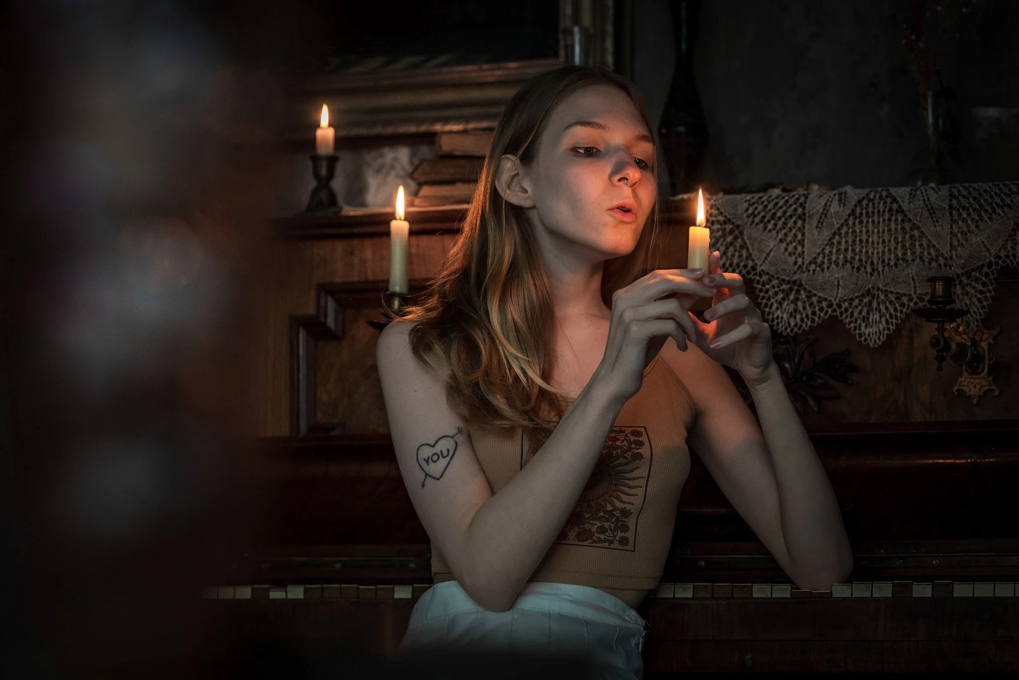 A young woman with long, wavy hair is sitting in a dimly lit room, holding a lit candle in her right hand near her face. She has a tattoo of a heart with the word "you" inside on her upper left arm. The background features a wooden wall, lit candles,