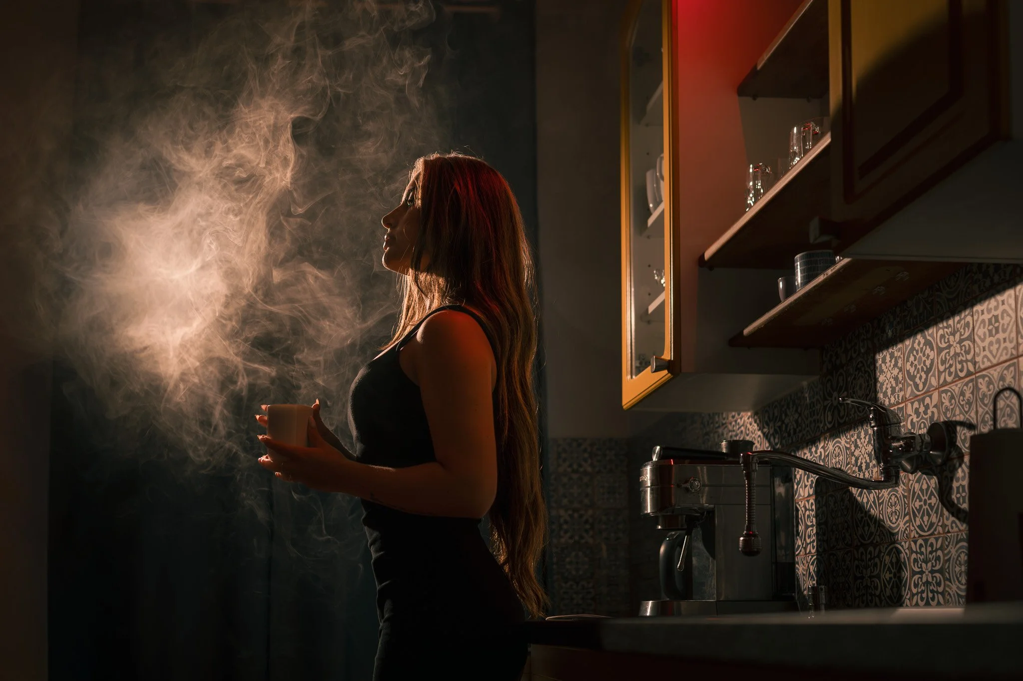Fotografie ženy s dlouhými vlasy stojící v tlumeně osvětlené kuchyni s hrnkem kávy v ruce, stoupající párou a vířícím kouřem nebo párou kolem hlavy.