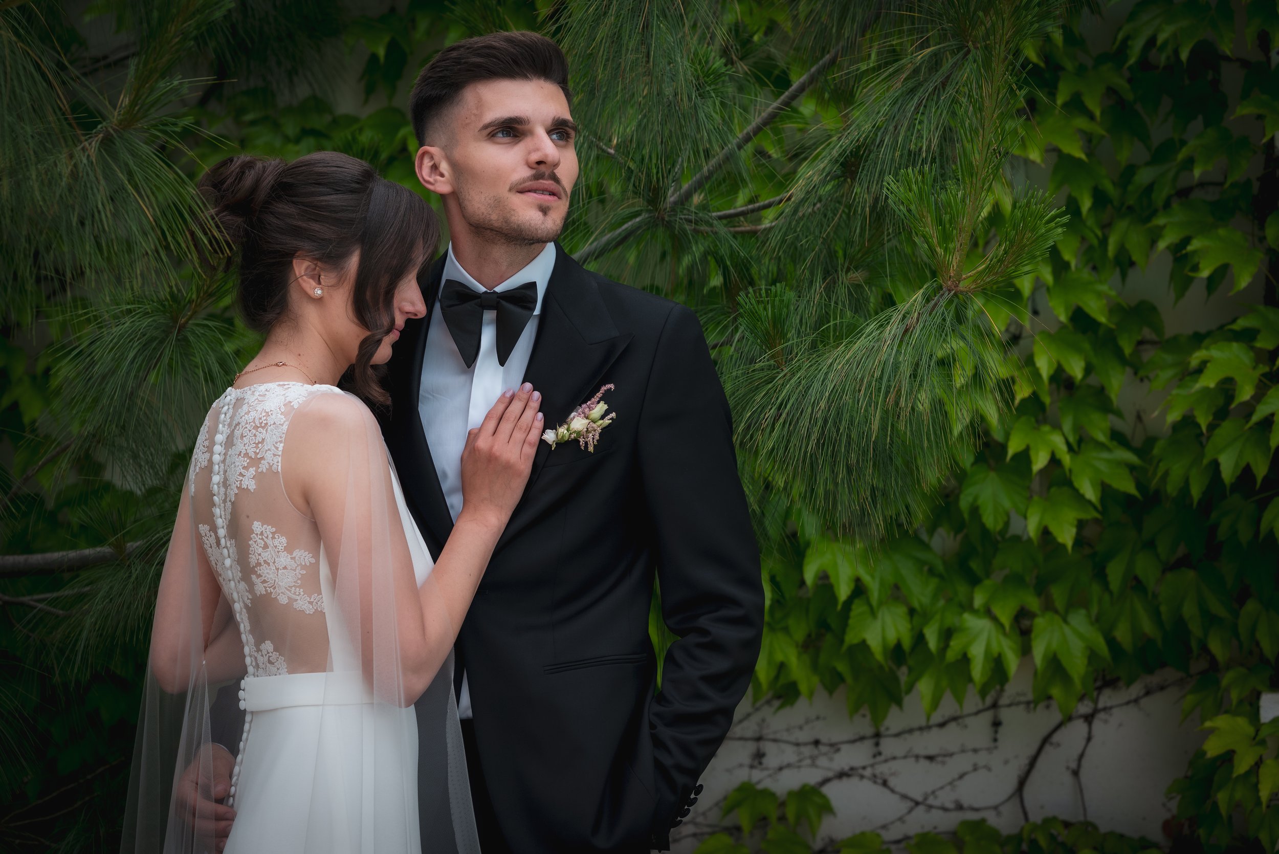 Bride in lace dress and groom in black tuxedo standing close, surrounded by green foliage.