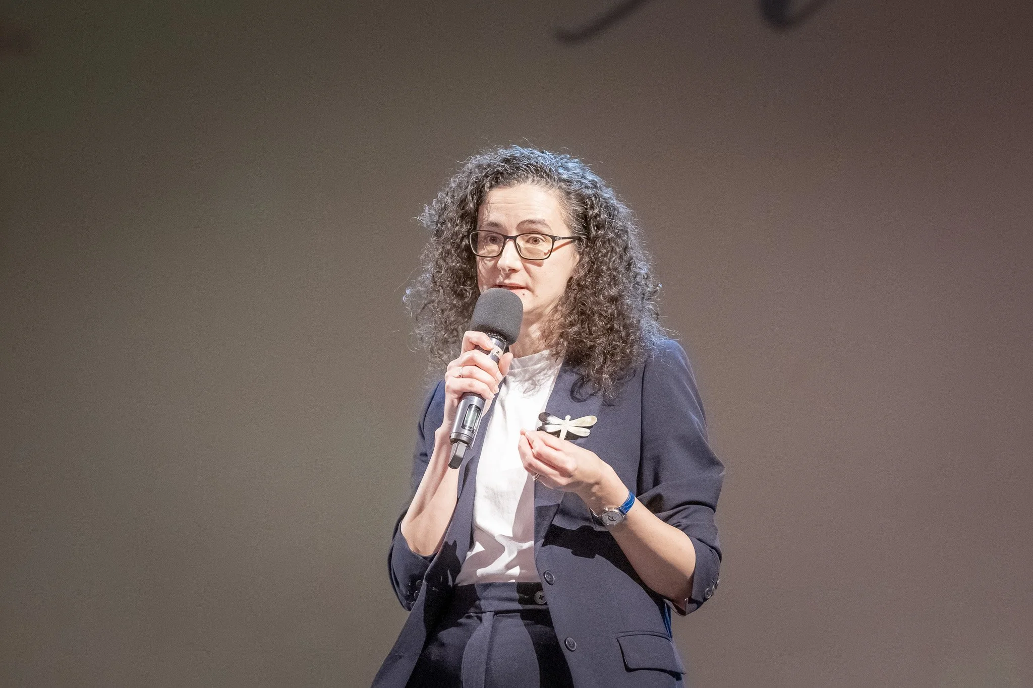 A woman with curly hair and glasses speaking into a microphone at an event, wearing a navy blazer and a white blouse.