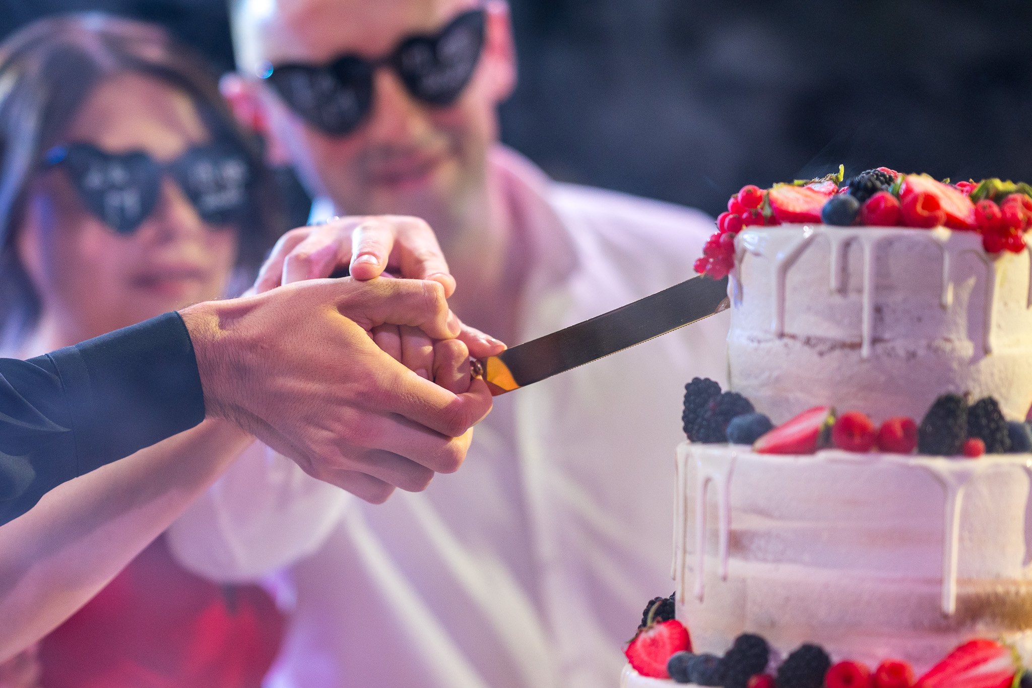 People wearing sunglasses cutting a wedding cake decorated with strawberries, blackberries, and redcurrants.