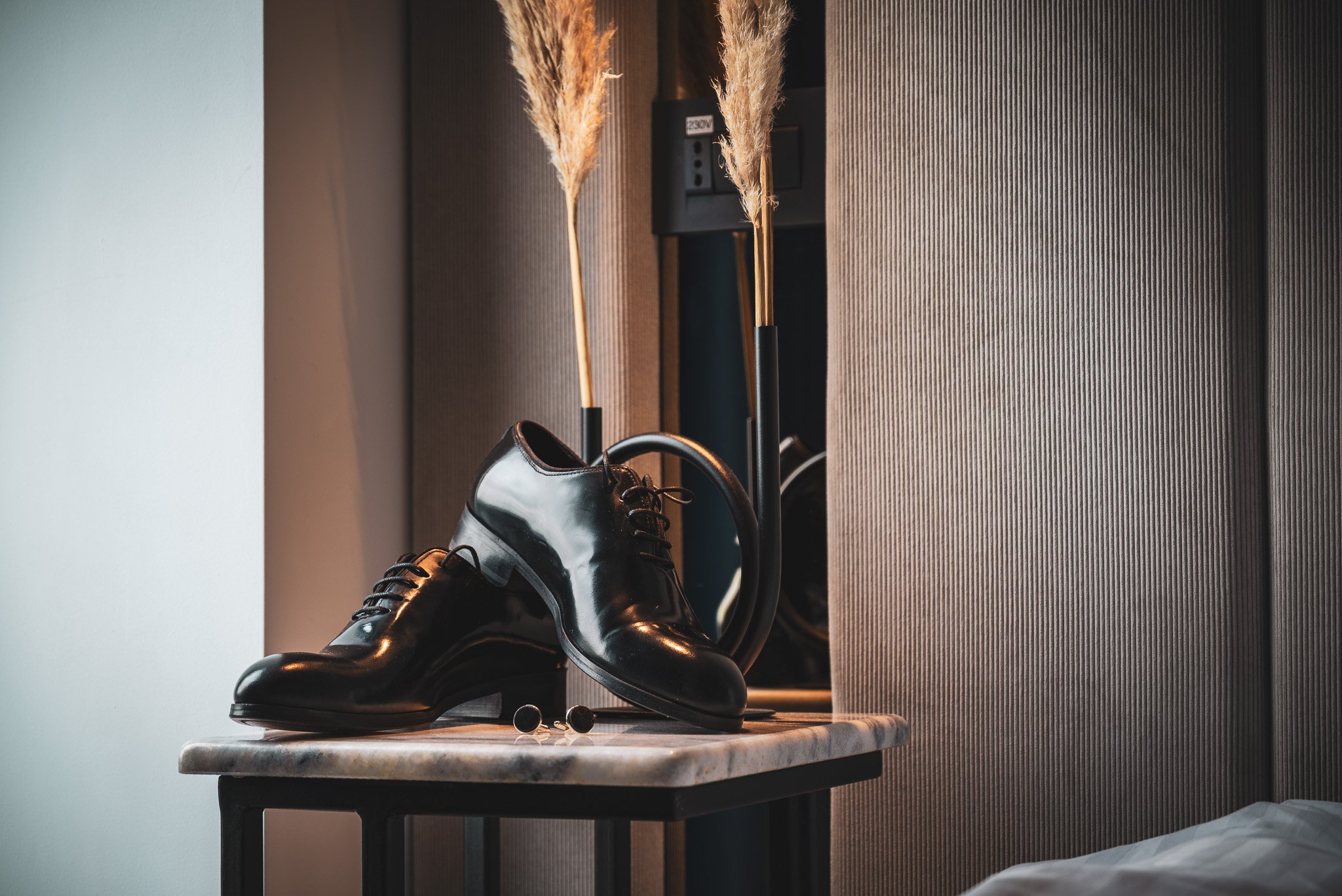Pár lesklých černých kožených společenských bot, náramkové hodinky a manžetové knoflíčky na odkládacím stolku s mramorovou deskou v moderní ložnici. Za stolkem jsou vysoké dekorativní suché traviny v černé váze a stěna s tmavými strukturovanými panel