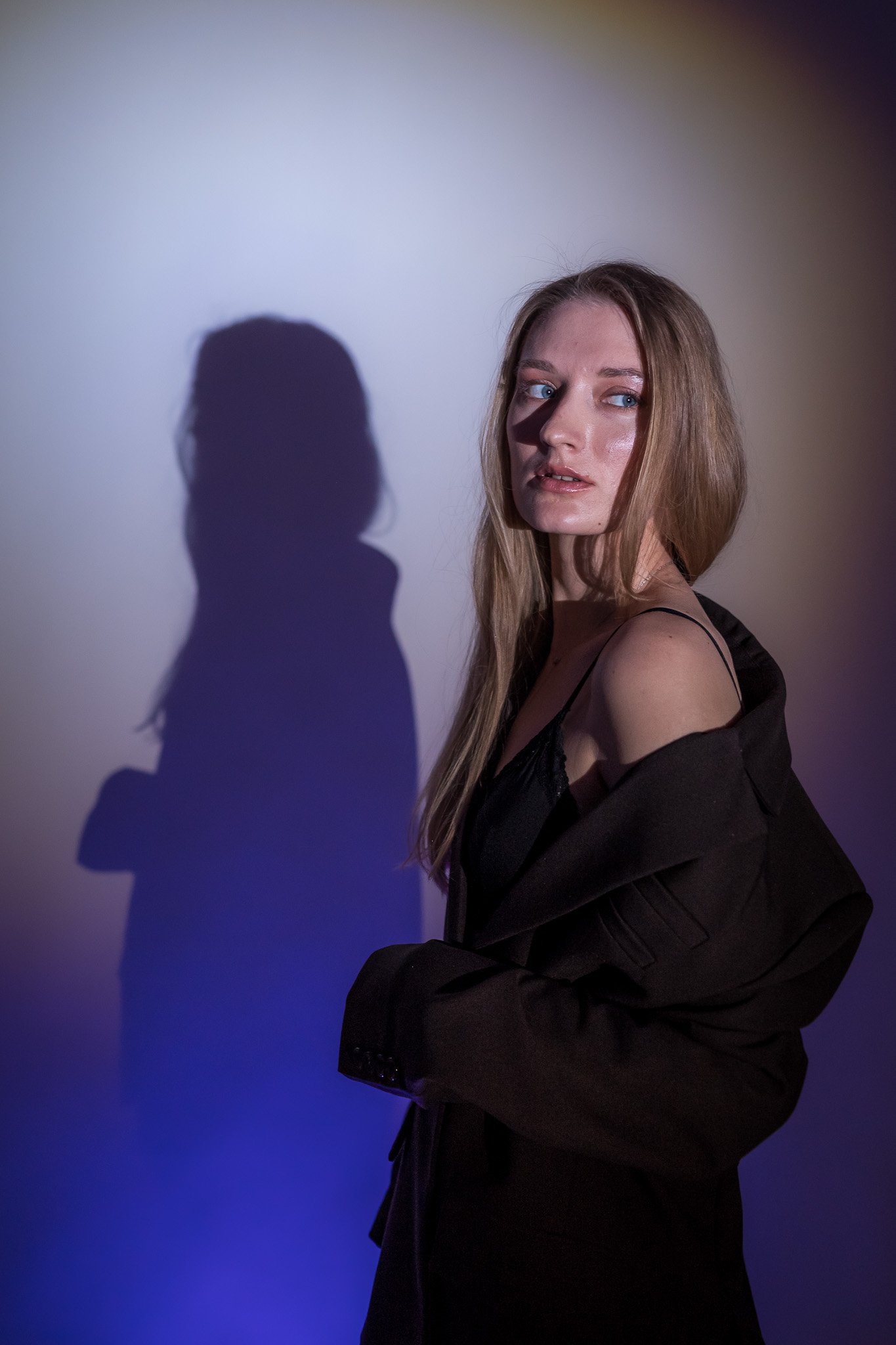 Woman in a black outfit with long hair posing against a gradient background with a shadow, looking to the side.