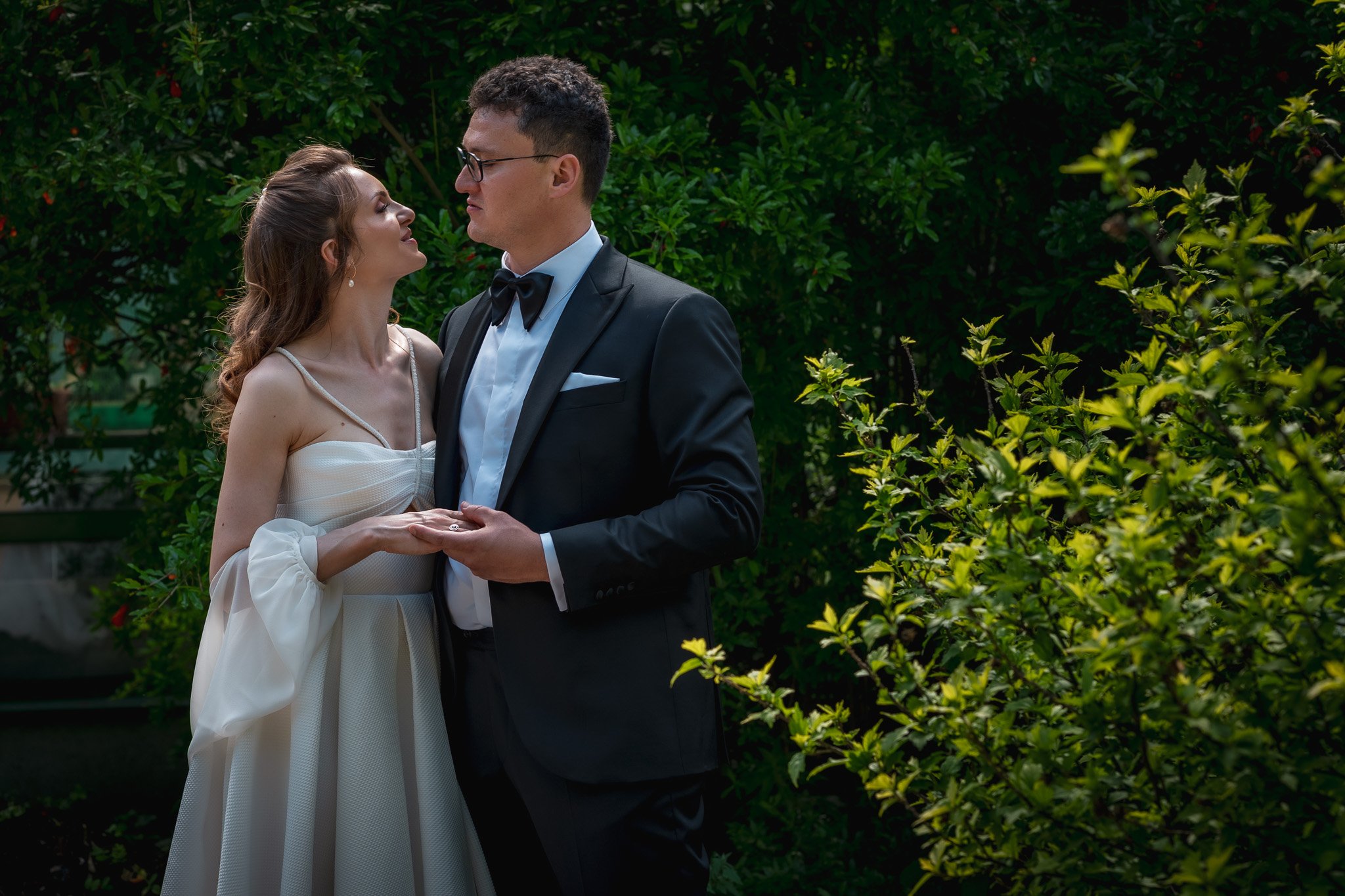 A couple in formal attire stands in a lush garden. The woman wears a white dress and the man wears a black tuxedo with a bow tie. They are holding hands and looking at each other.