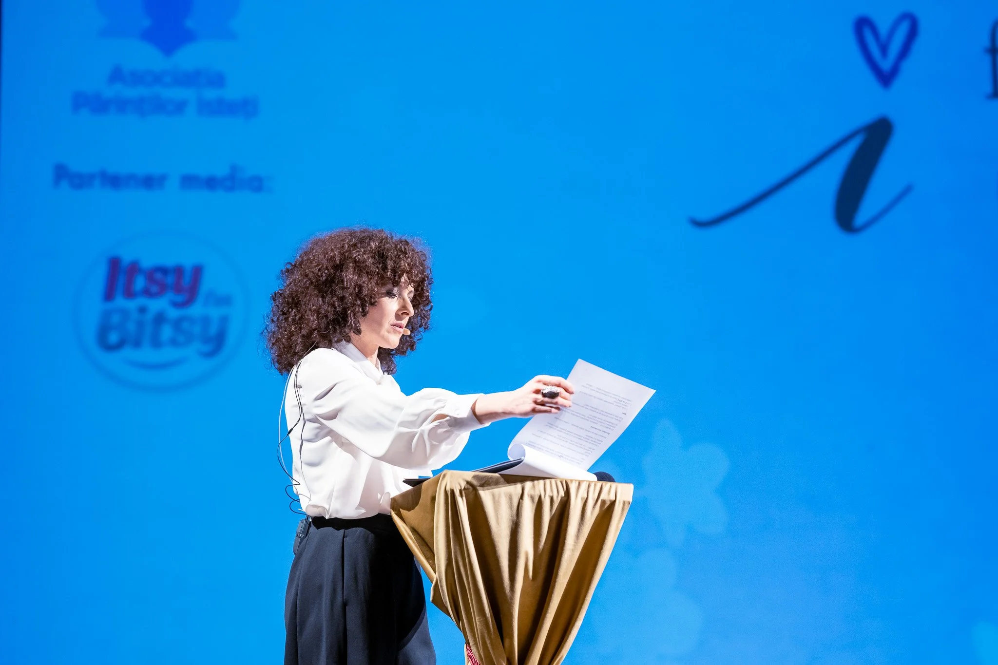 A woman with curly hair, wearing a white shirt and black pants, is reading a document on a podium draped with a gold-colored cloth during a presentation or speech. The background features a blue screen with the "Itsy Bitsy" logo and blue text.
