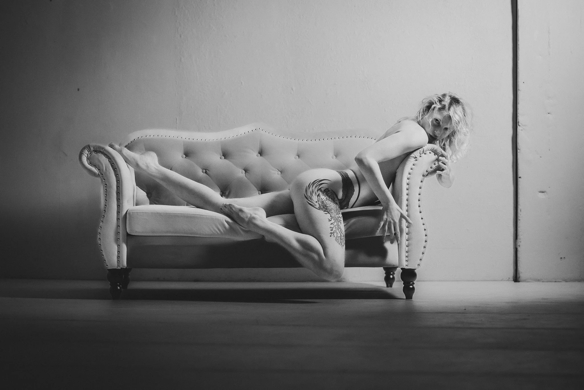 Black and white photo of a person with a tattoo, lounging provocatively on a vintage sofa.