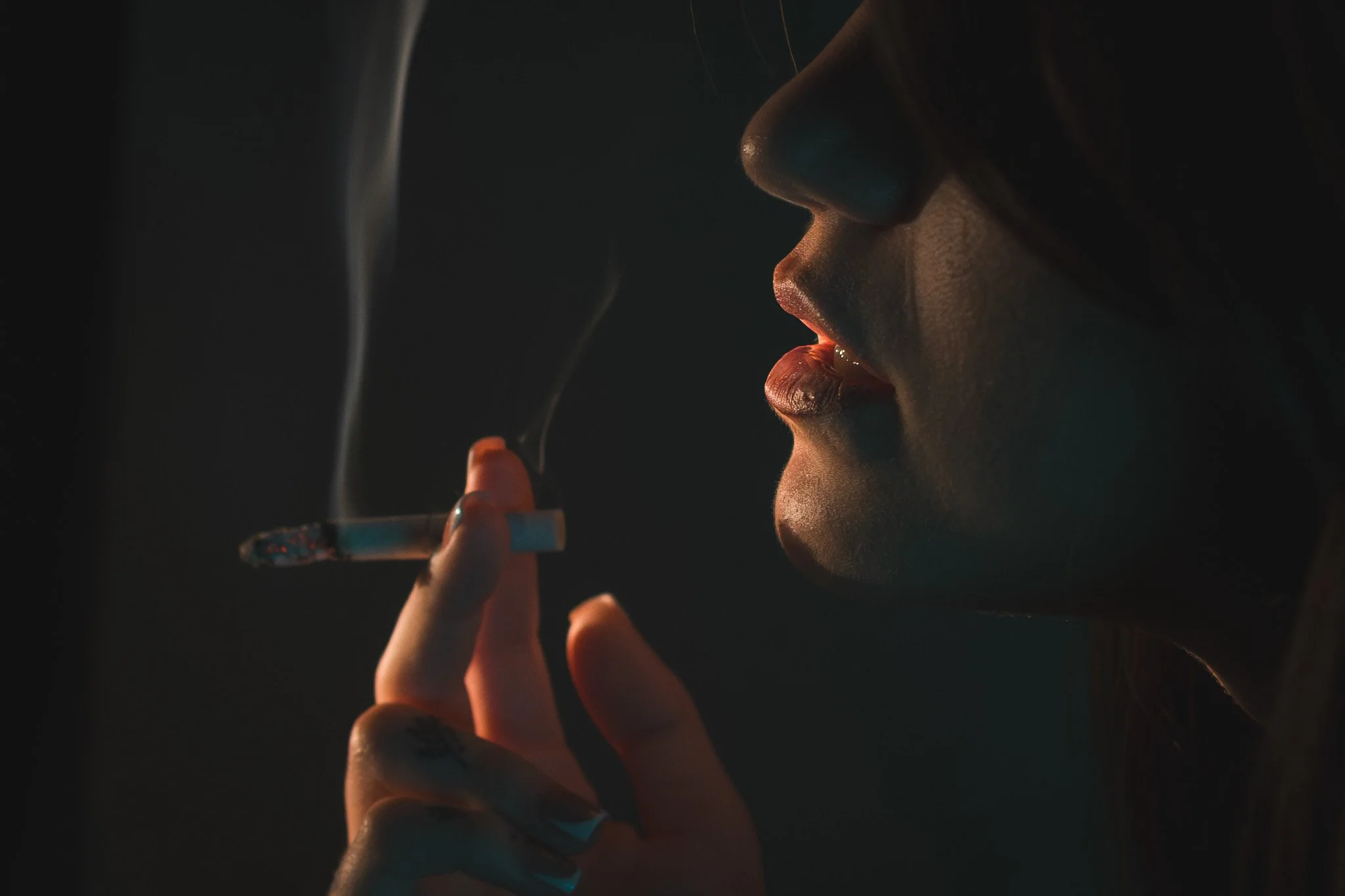 A close-up photograph of a woman’s face in profile, holding a lit cigarette to her lips, with smoke curling upward in the dim light.