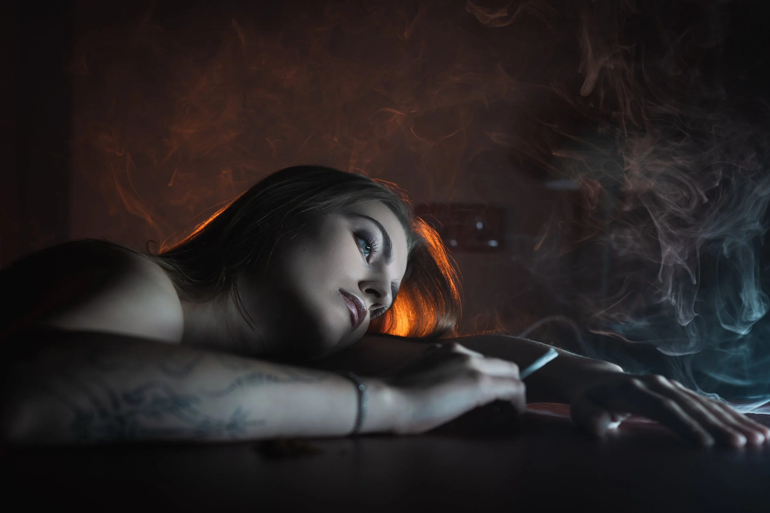 Fotografie ženy s hnědými vlasy a modrýma očima, ležící obklopená kouřem, osvětlená oranžovým a modrým světlem.