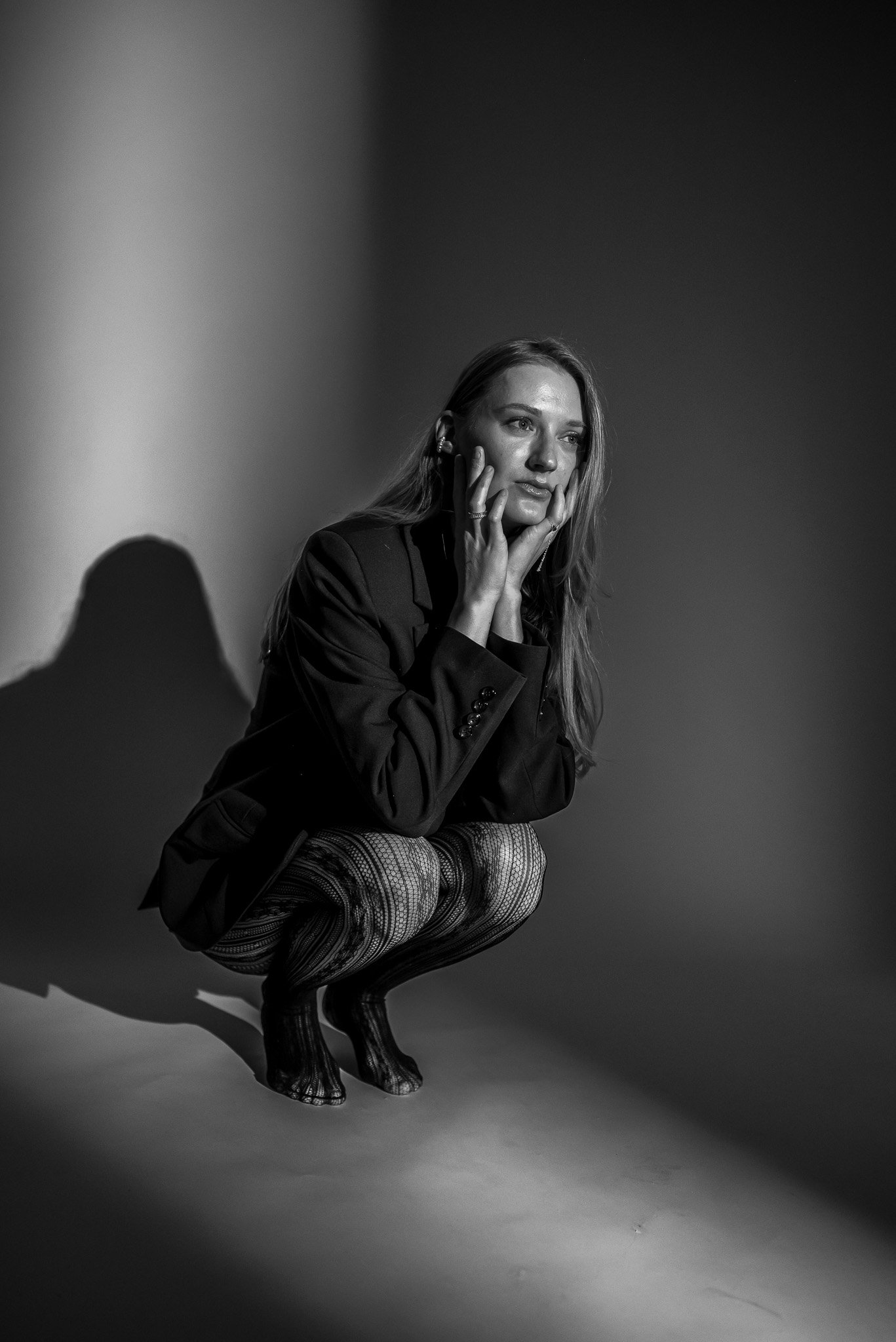 Black and white photo of a woman crouching, wearing a blazer and textured leggings, with her hands on her face, set against a subtle gradient background.