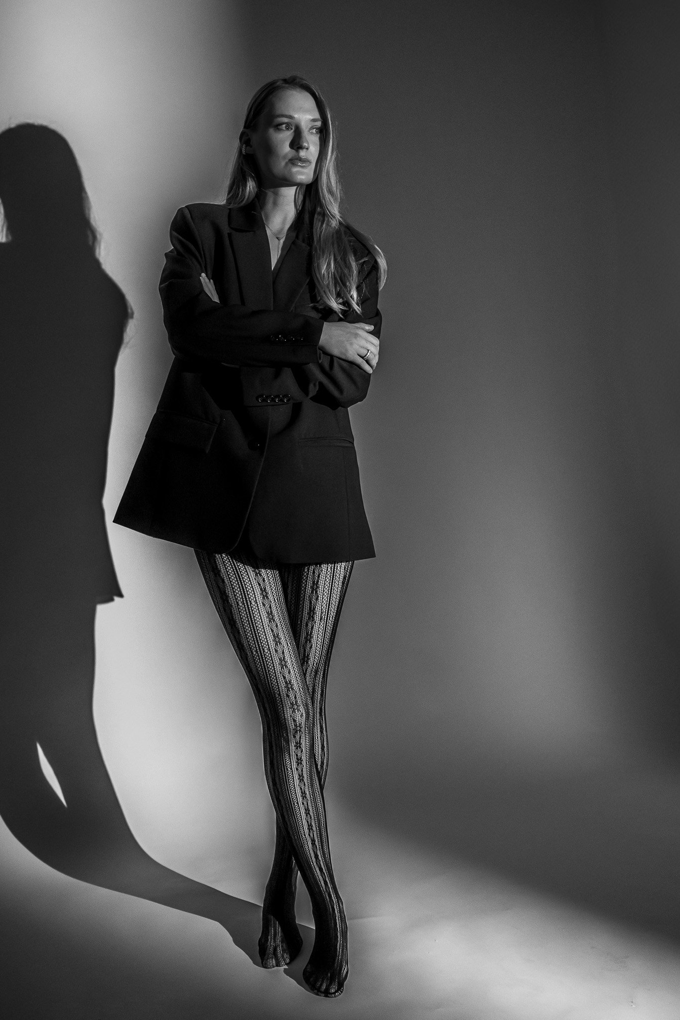 Black and white photo of woman in blazer and patterned tights standing with arms crossed, casting a shadow on the wall.