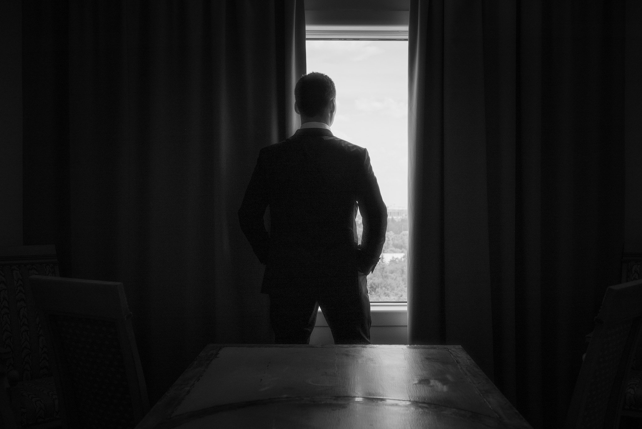 Silhouette of a man in a suit standing at a window, looking outside, with curtains partially drawn.
