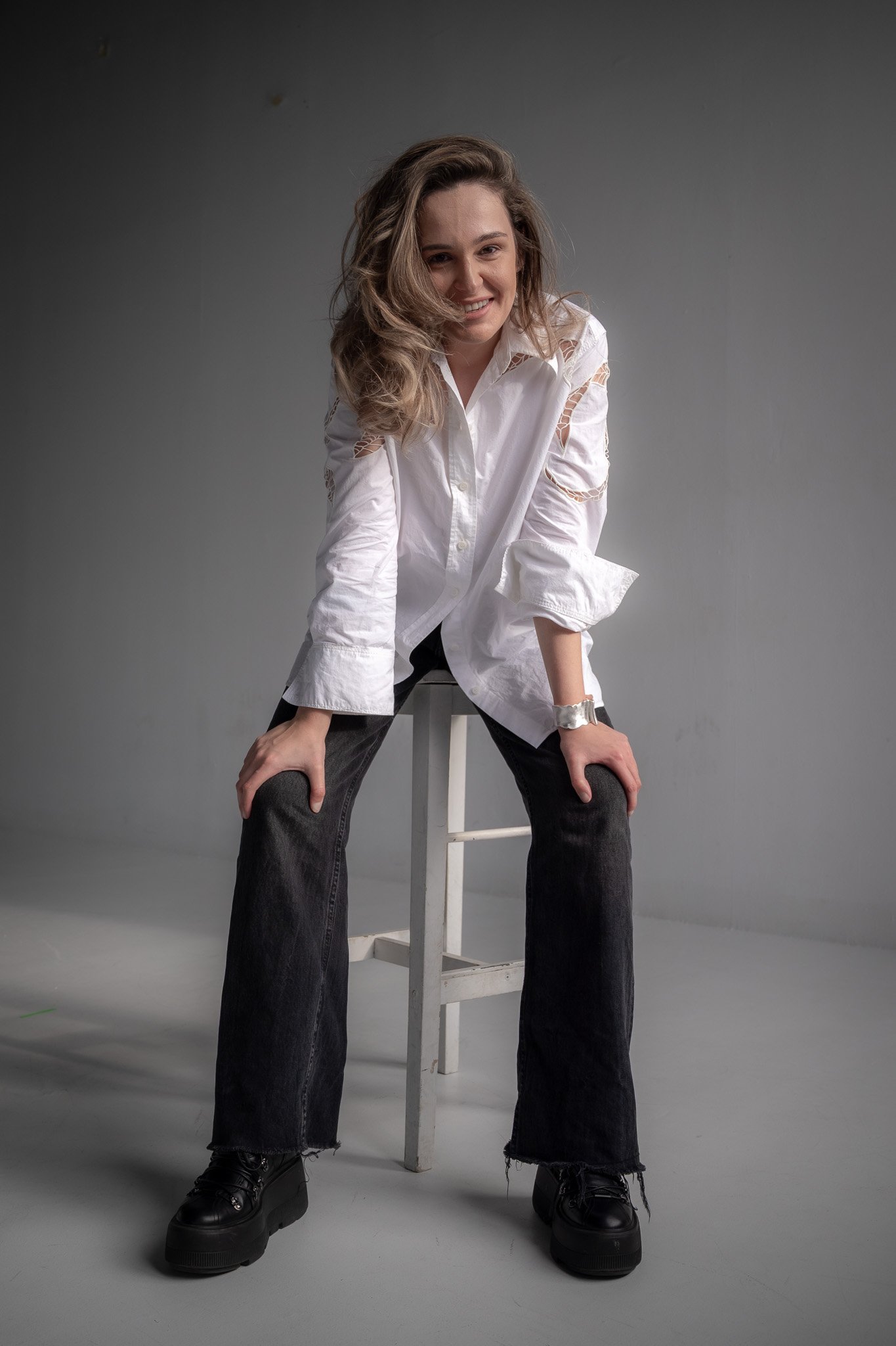 Person sitting on a stool wearing a white shirt and black pants, smiling, in a gray room.