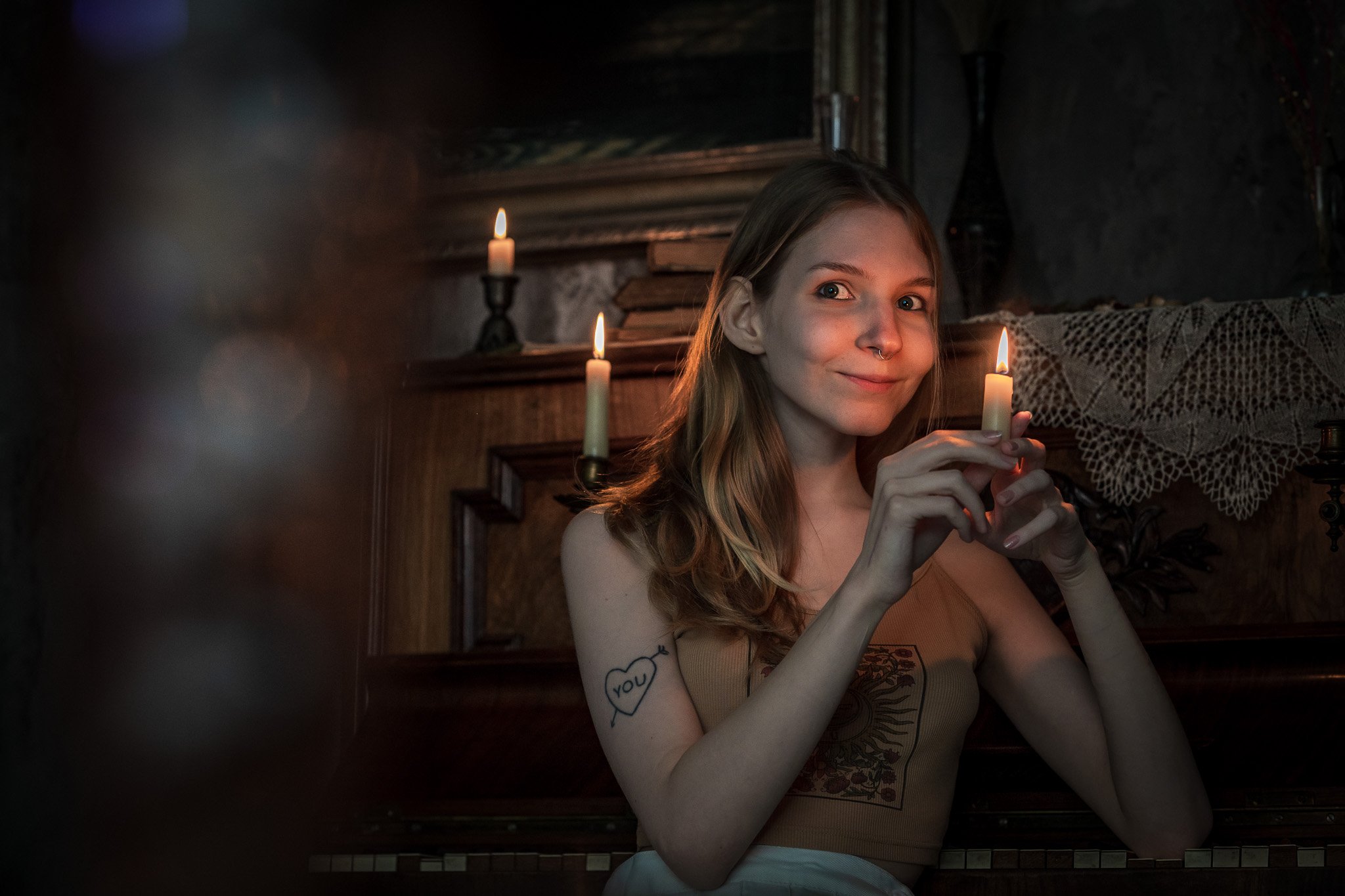 A young woman with long red hair and a septum piercing holding a lit candle, smiling softly in a dimly lit room with wooden furniture and candles.
