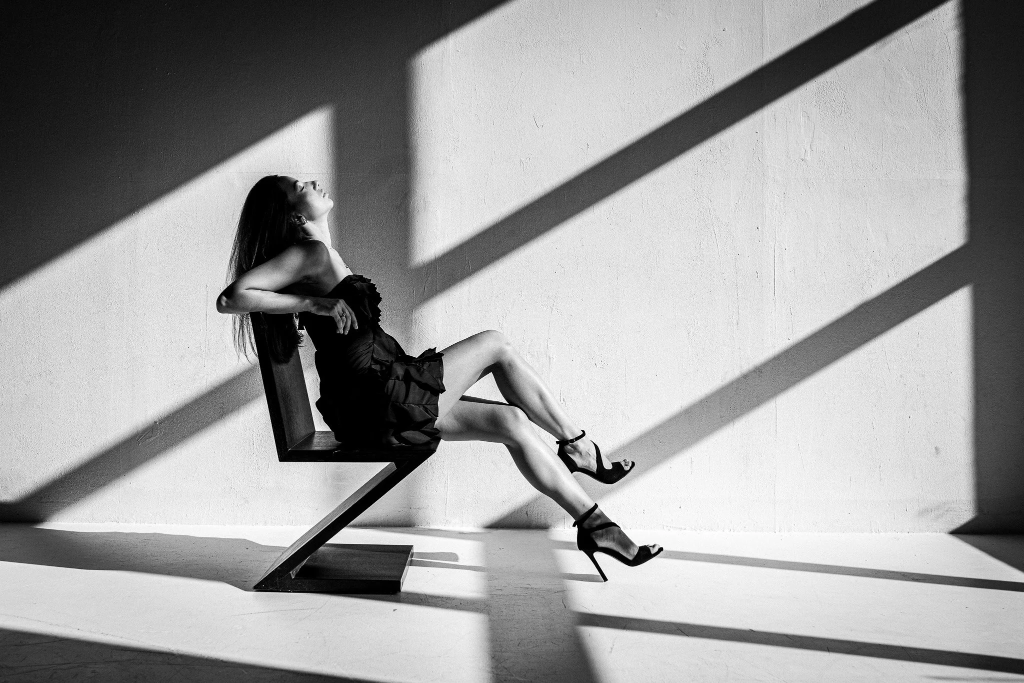 A woman in a black dress and high heels sitting on a modern chair, leaning back with her face tilted upward, with dramatic shadows cast on the wall behind her in a monochrome setting.