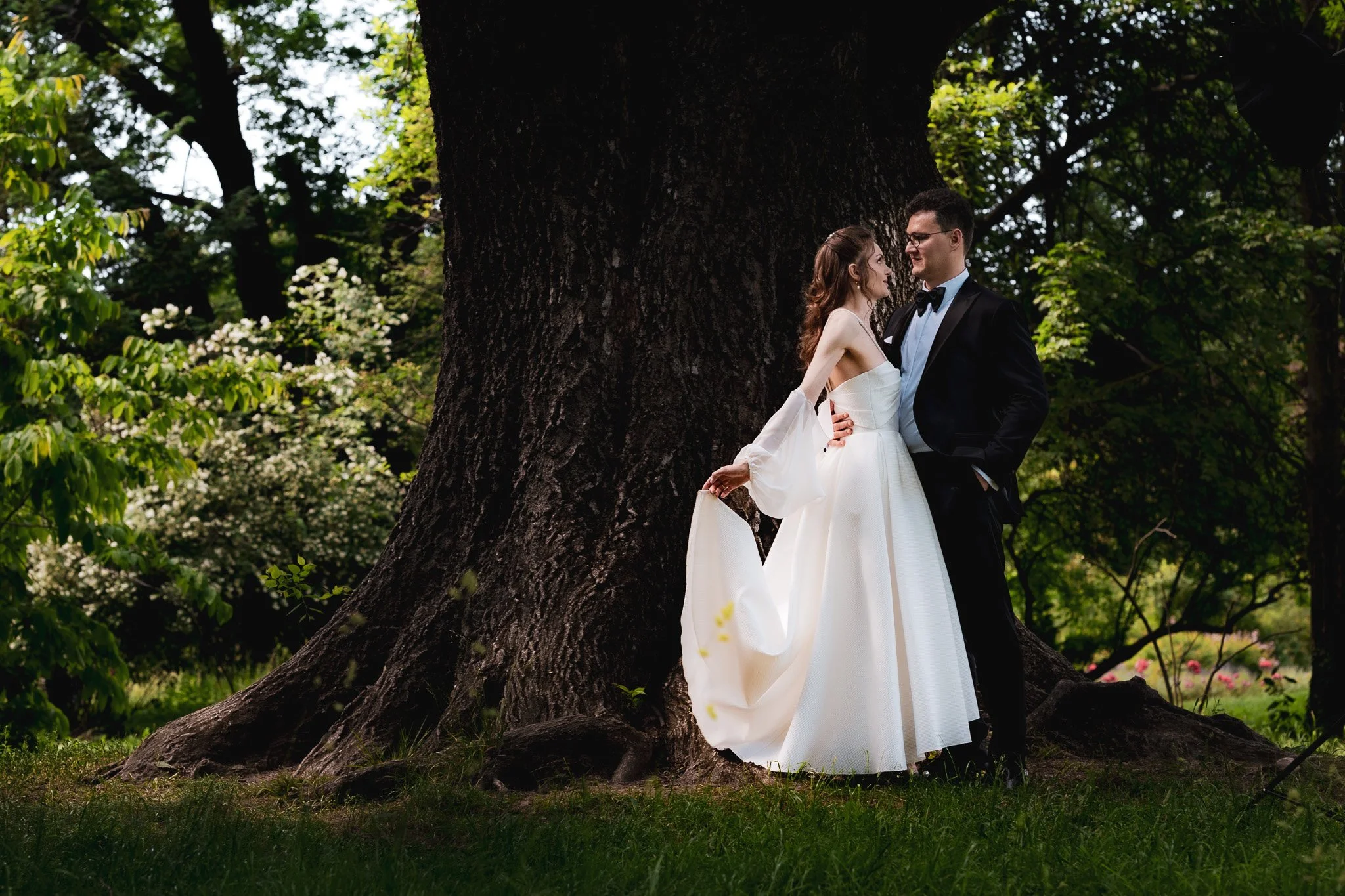 A bride and groom standing close together under a large tree in a green garden, gazing at each other. The bride is in a white wedding dress, holding its flowing train, and the groom is in a black tuxedo with a bow tie. Sunlight filters through the le