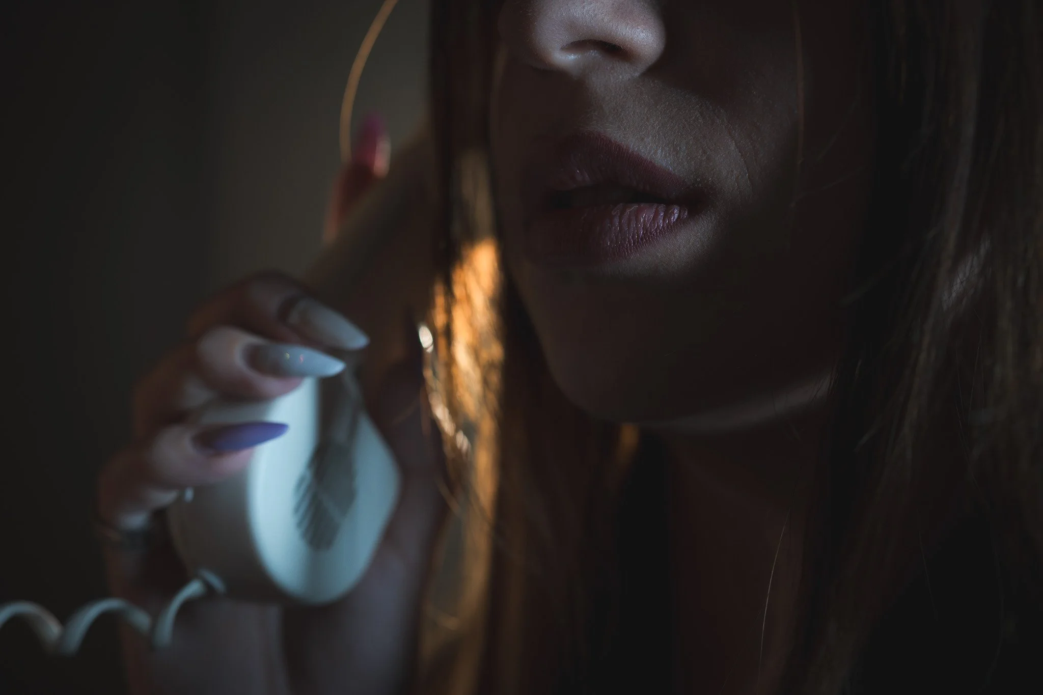 A close-up of a woman with painted nails and earrings, talking on a landline phone in dim lighting.