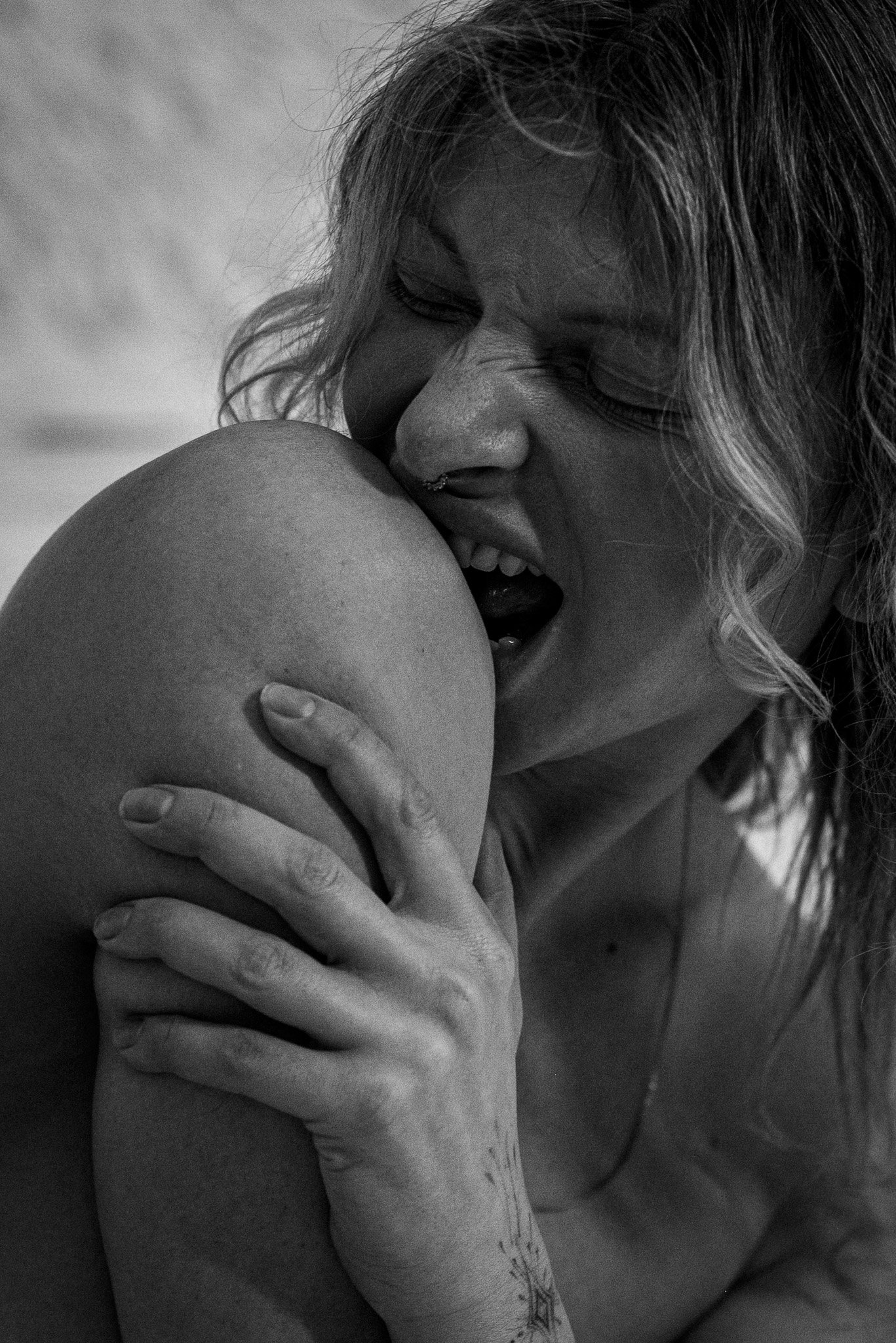 Black and white image of a person biting their own knee while holding it with one hand.