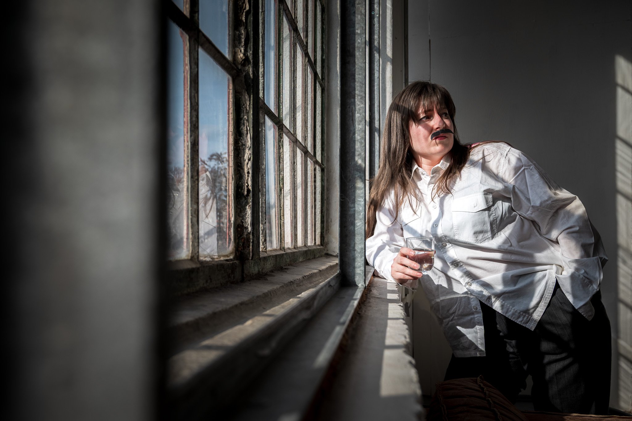 Person wearing a white shirt and fake mustache holds a glass of water, leaning against a window with sunlight streaming in.
